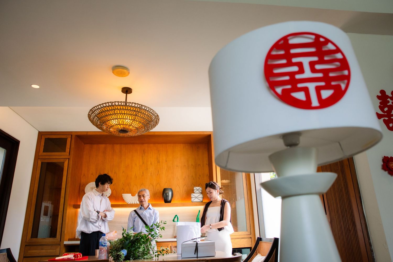 Two people stand near a wooden table with flowers in a warm, modern interior, with a Chinese double happiness lamp in the foreground.