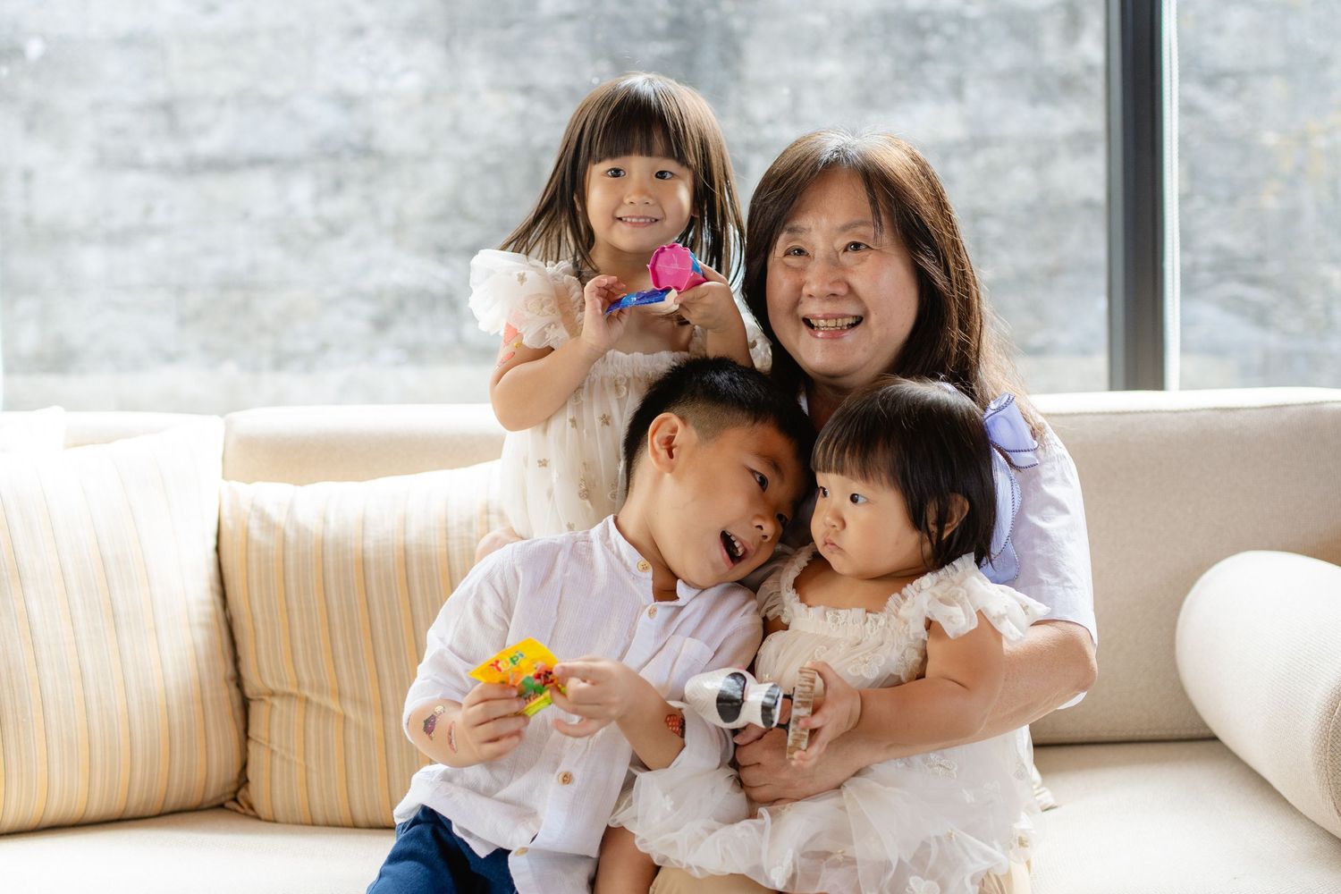 Smiling grandmother hugging three young children on a white sofa near a bright window in a cozy living room.