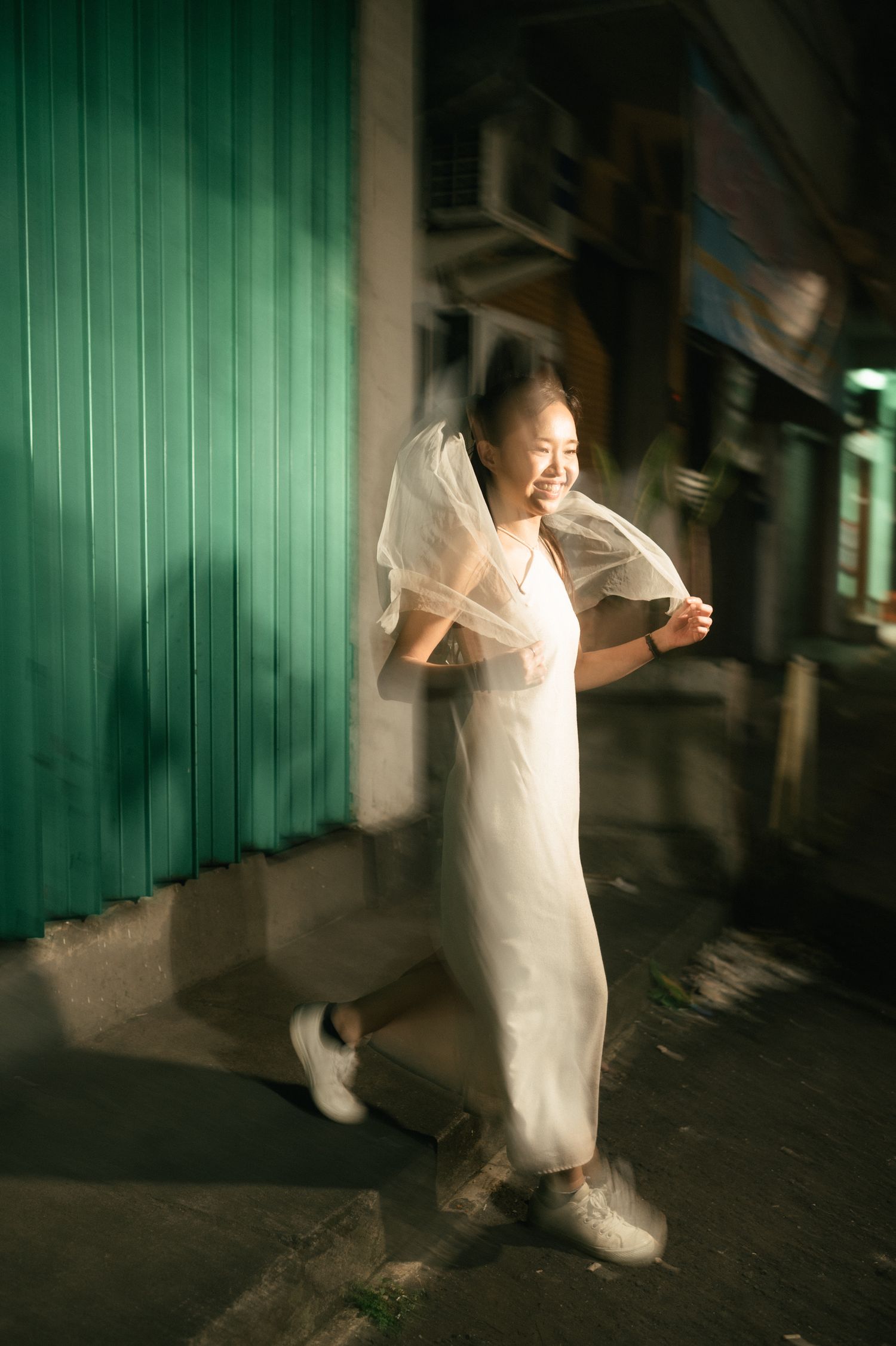 Smiling woman in white dress and veil walking outdoors near a teal wall in soft evening light.
