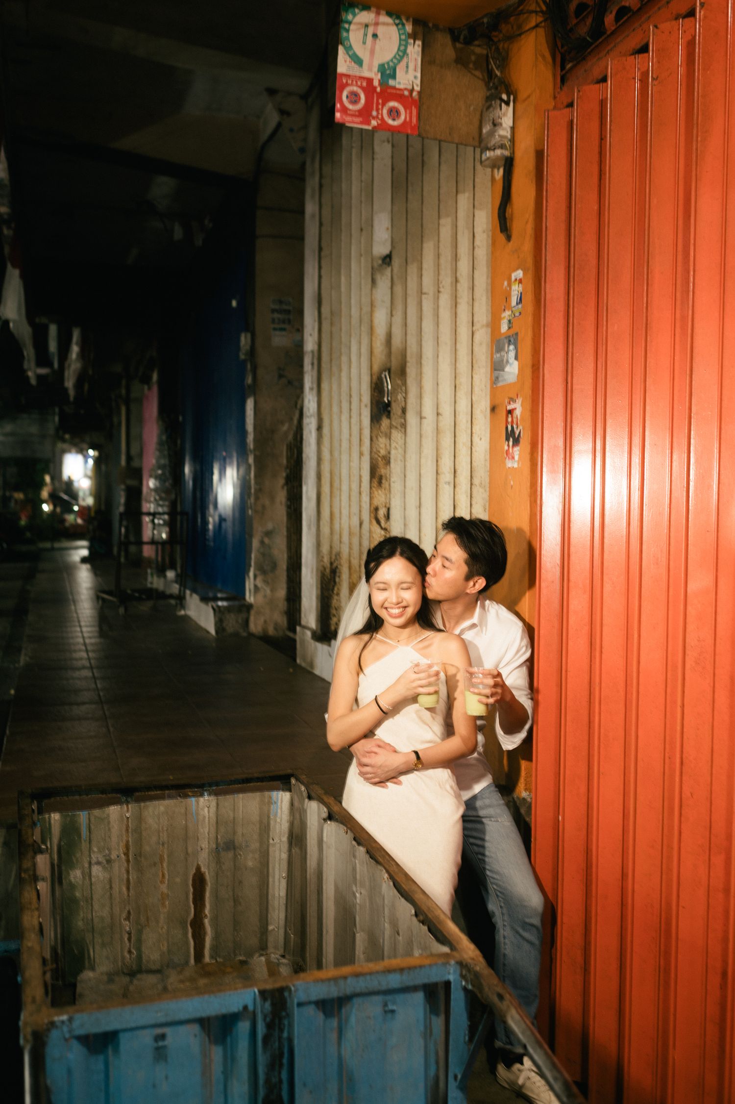 Couple sharing a romantic moment in a night alley, man kissing woman holding drinks by colorful shipping containers.