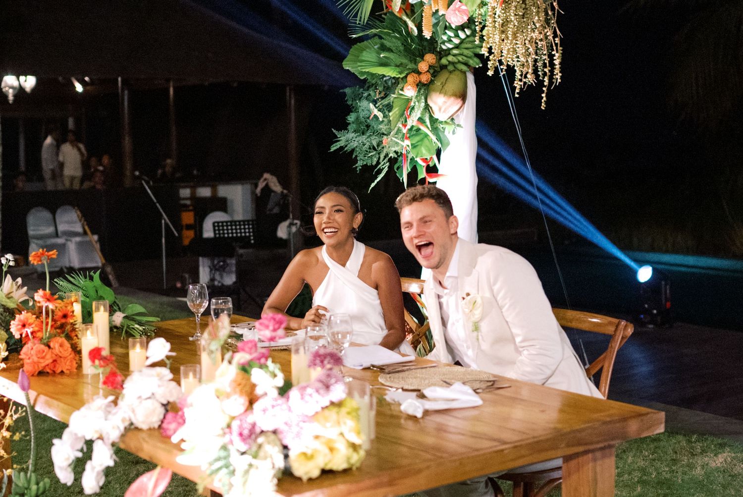 Bride and groom laughing joyfully at wedding reception table with tropical floral arrangements at night.