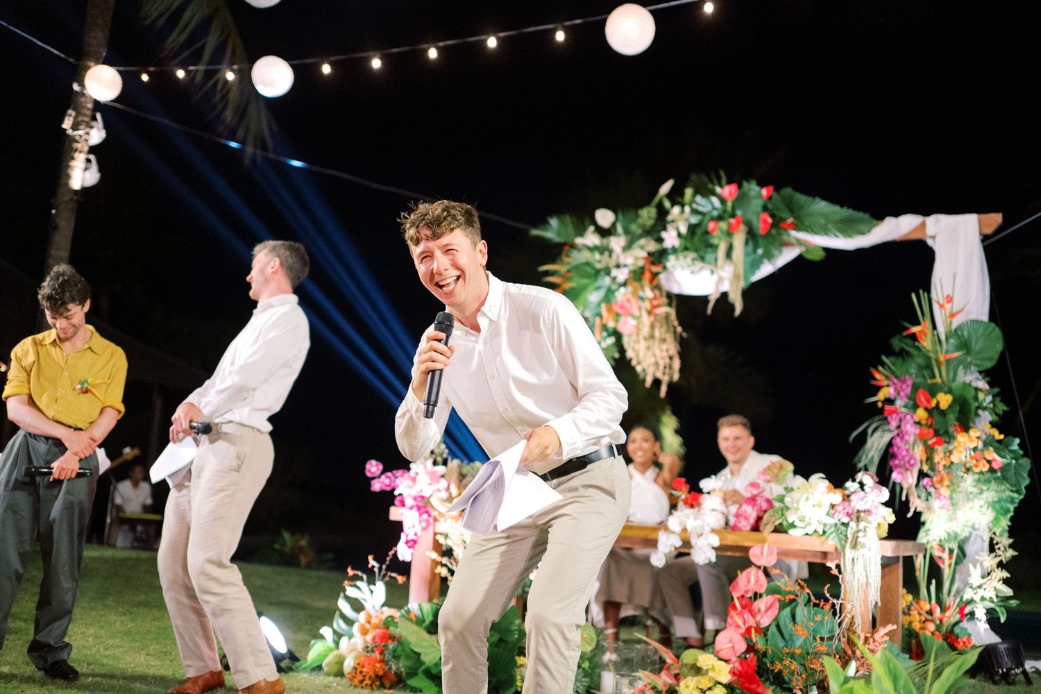 Man with microphone performing at tropical outdoor wedding reception with floral arrangements and string lights.