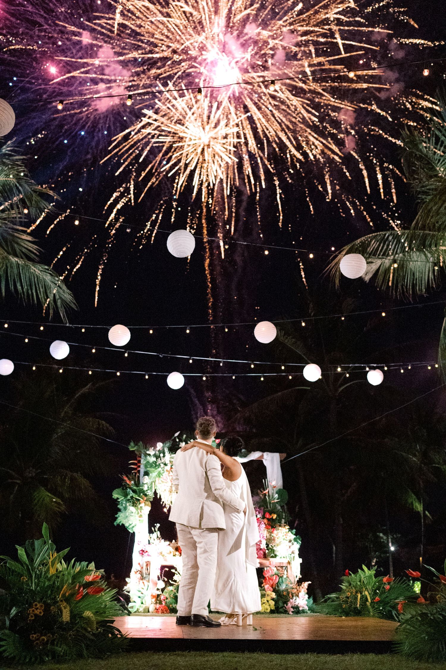 Couple sharing first dance under spectacular fireworks display at tropical outdoor wedding reception with string lights.