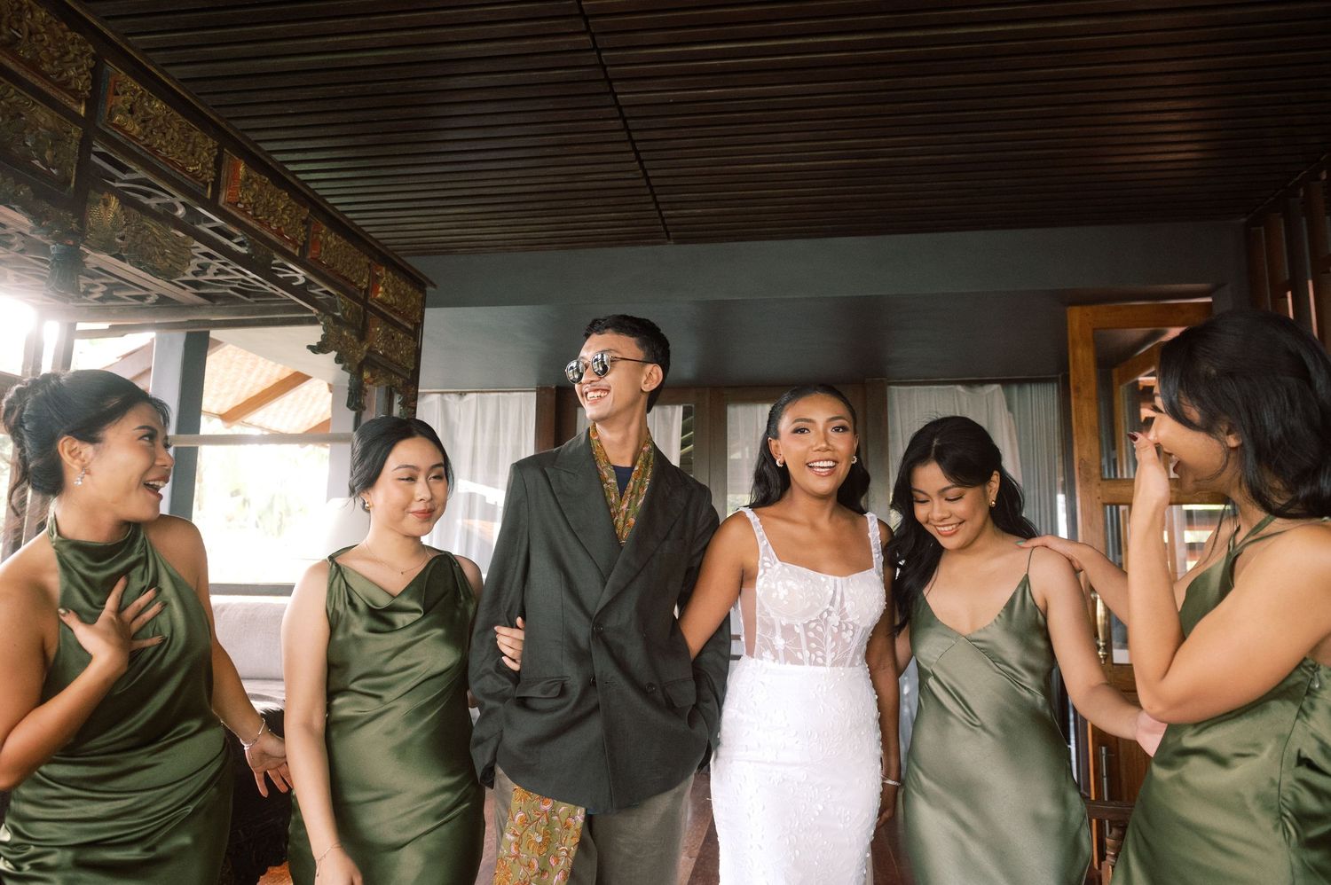 Bride in white lace gown with bridesmaids in olive green satin dresses and groomsman in dark blazer.