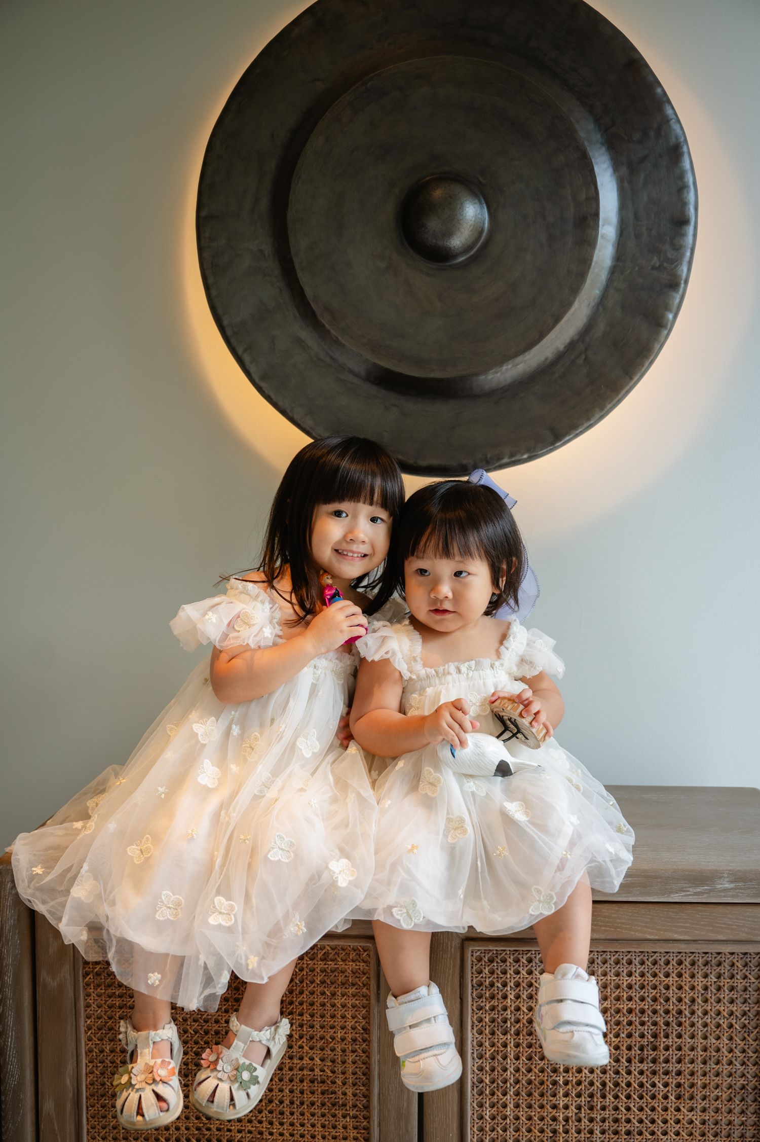 Two young girls in matching white tulle dresses sit together in front of a decorative gong wall art.