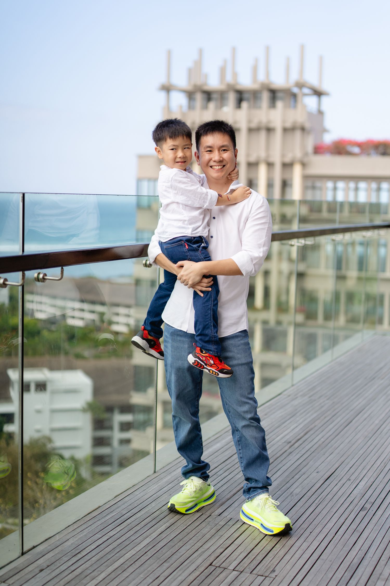 A smiling Asian man holds a young boy on a rooftop terrace with city buildings and blue sky in the background.