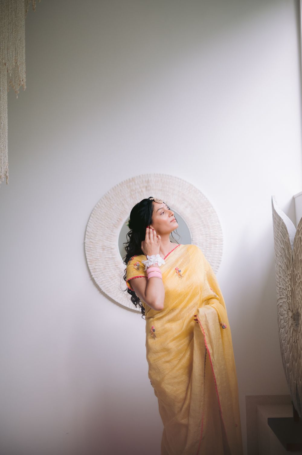 Woman in yellow saree posing gracefully in front of a round woven wall mirror in a serene interior.