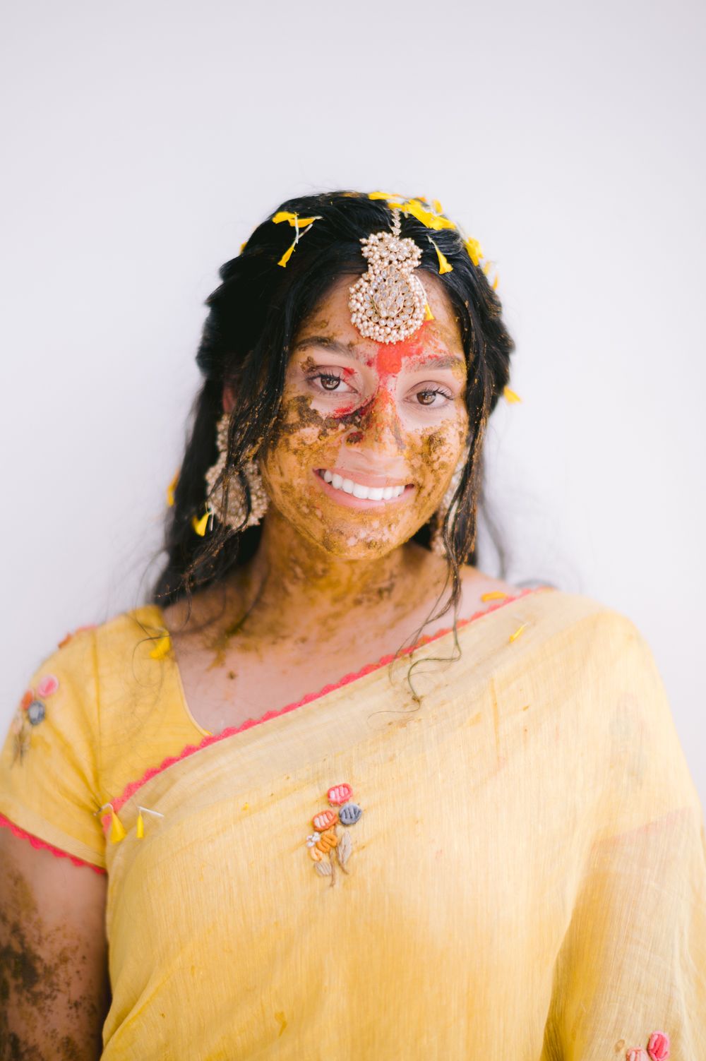 Smiling Indian bride in yellow saree with haldi paste on face, wearing maang tikka and earrings during haldi ceremony.