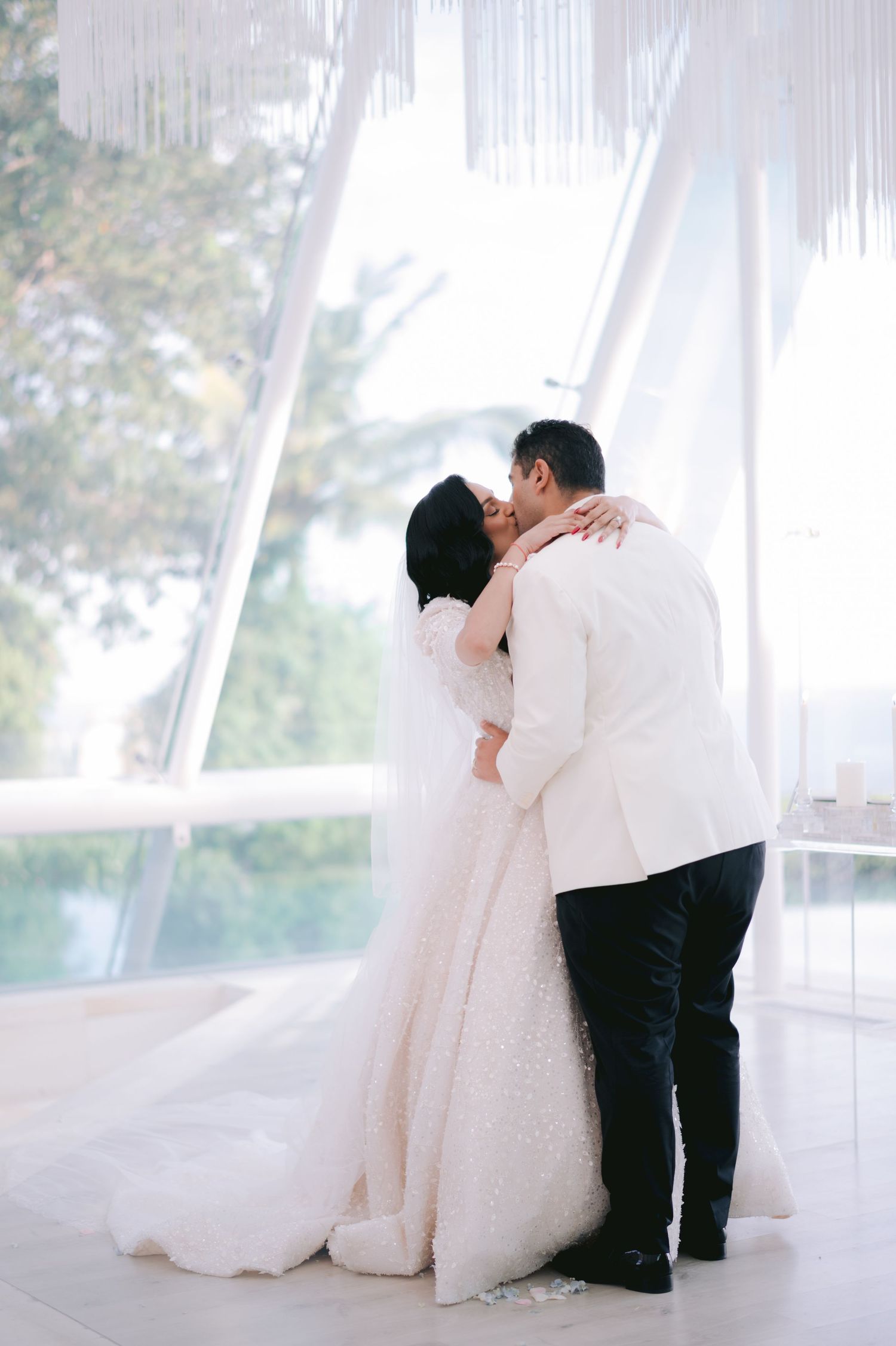 Bride and groom share first kiss in elegant white wedding venue with flowing drapes and floor-to-ceiling windows.