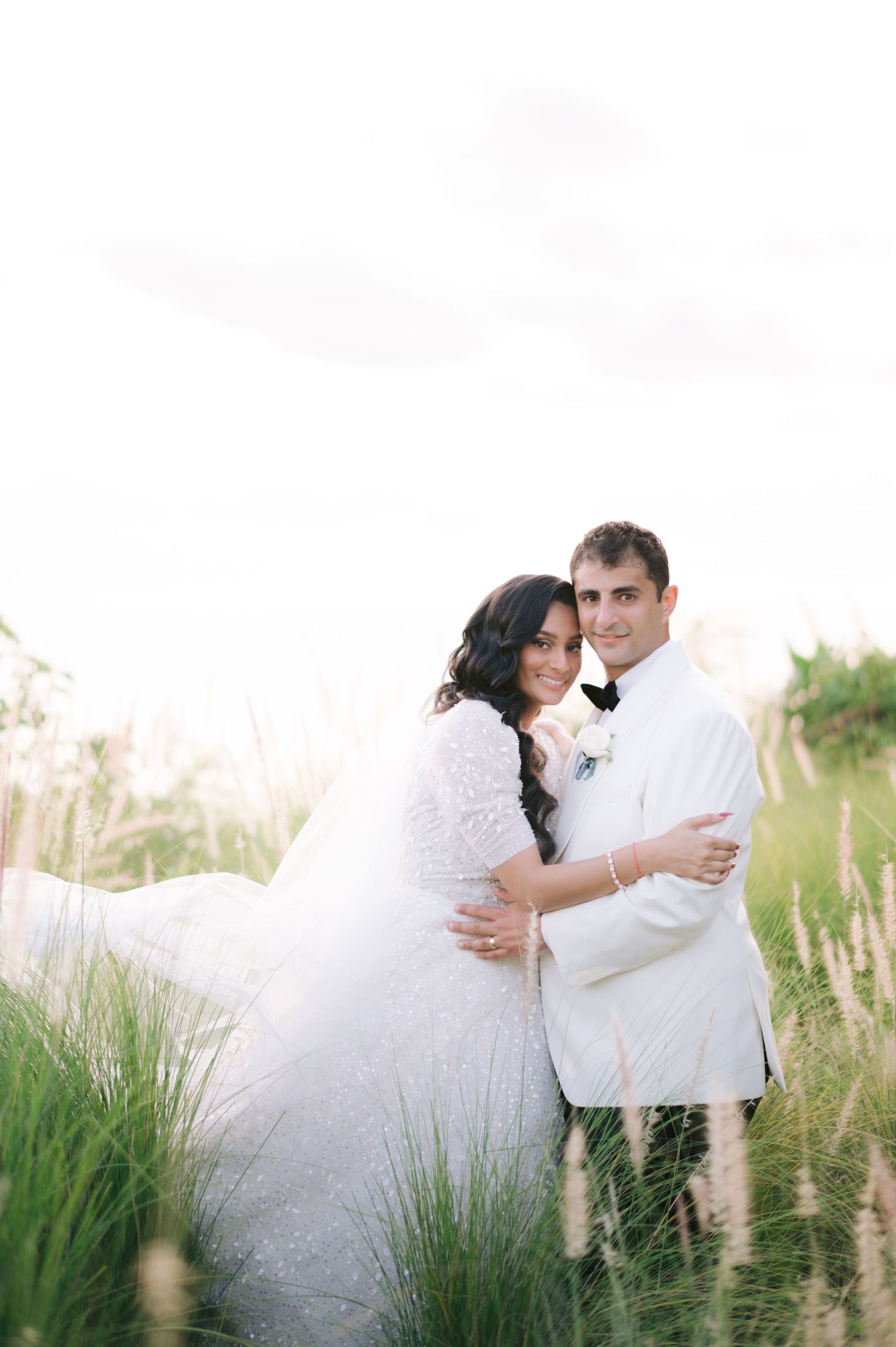 A bride and groom embrace in a lush green field, wearing white wedding attire, posing for a romantic outdoor portrait.