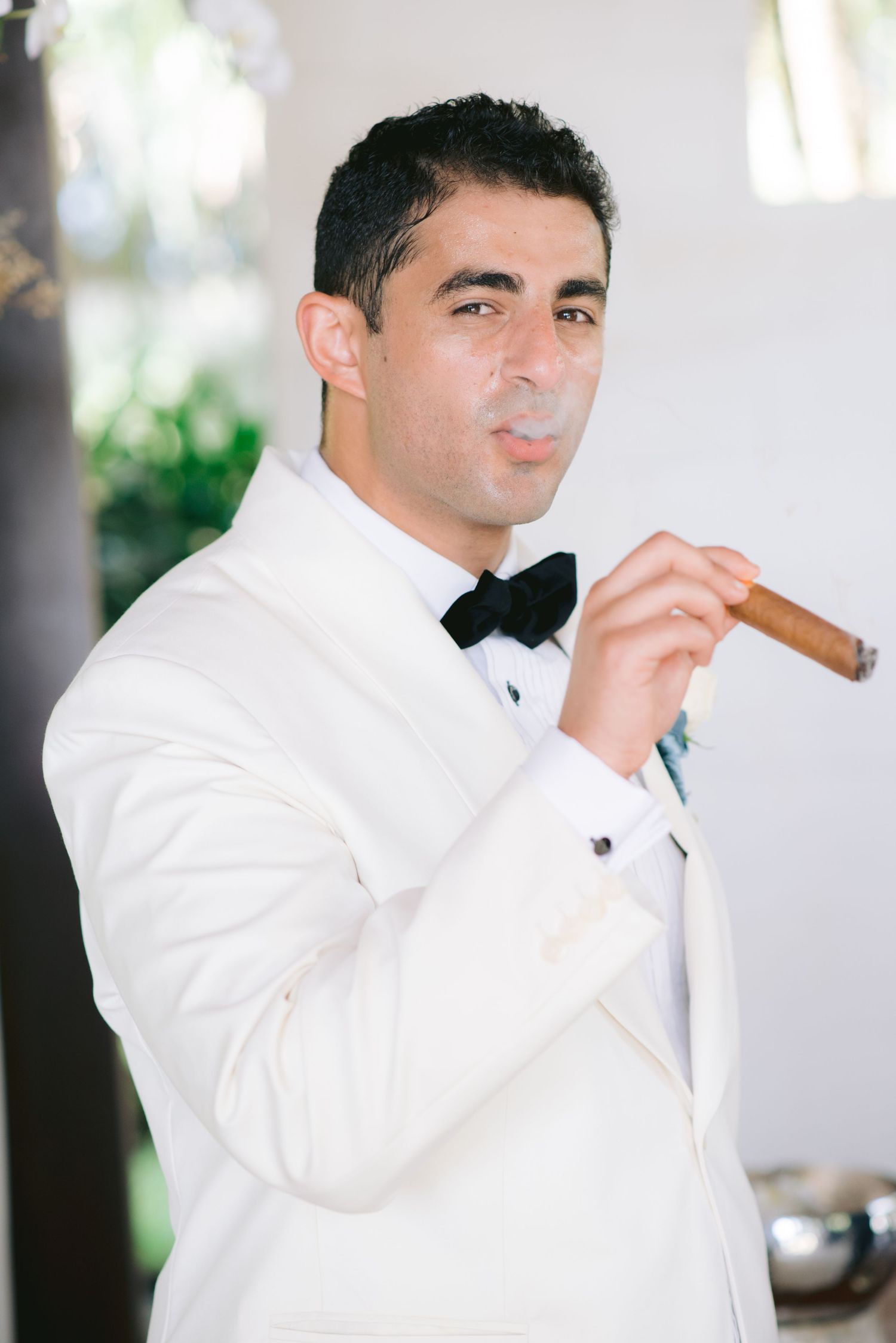 Man in white tuxedo and black bow tie smoking a cigar at a formal event.