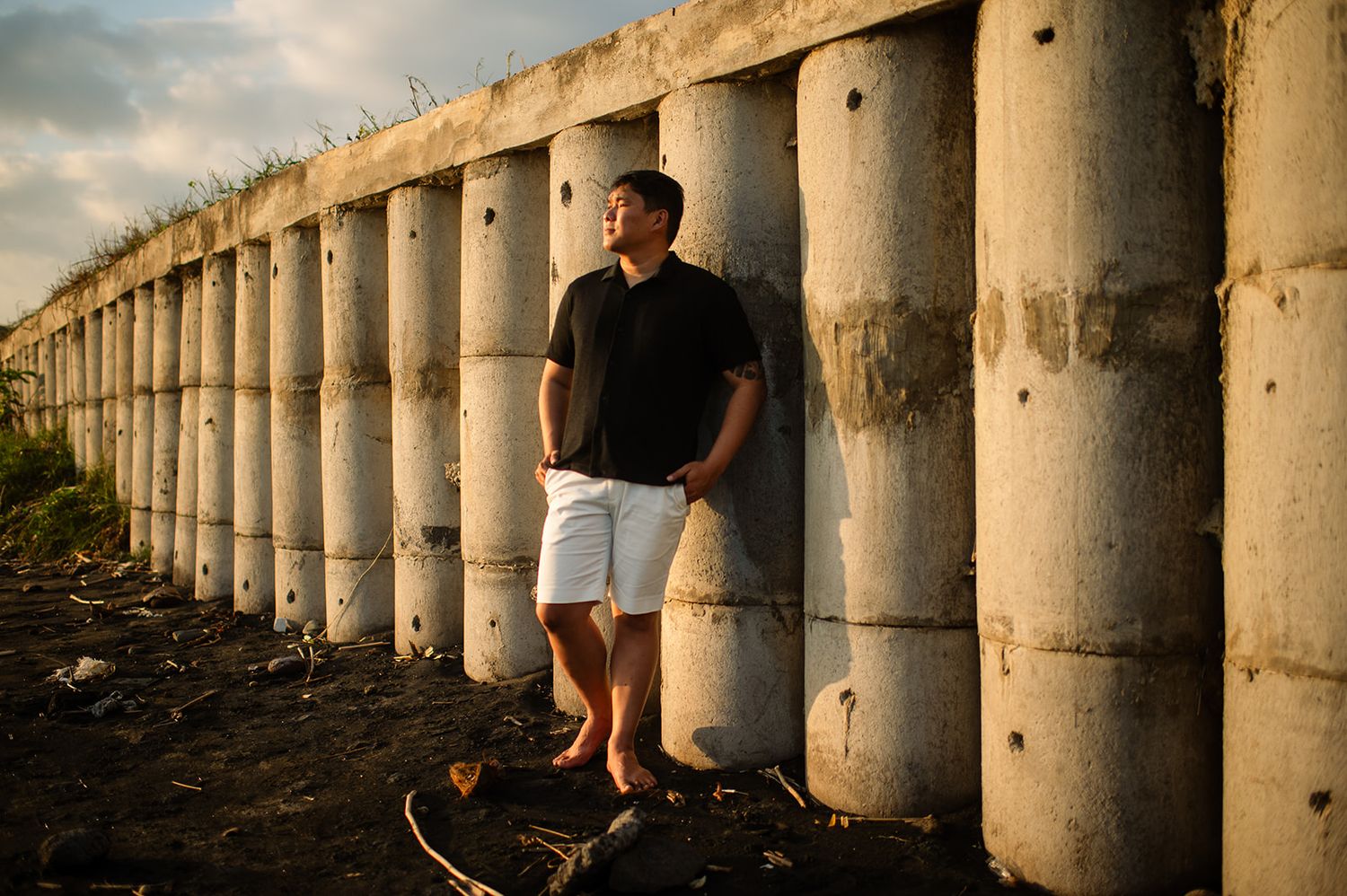 Young man in black shirt and white shorts leaning against concrete cylinders at sunset.