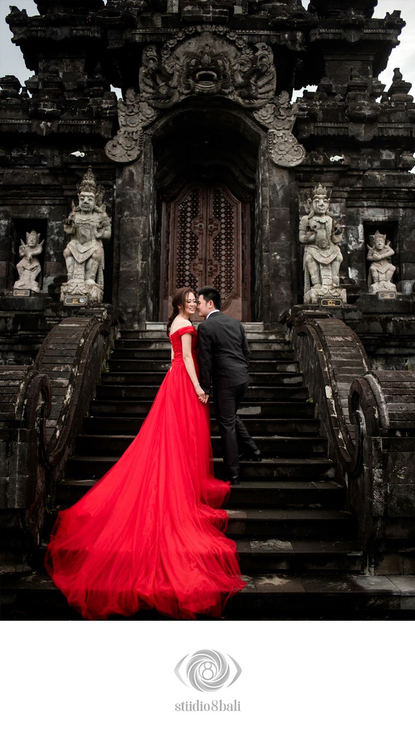 Couple in red gown and black tuxedo posing on ancient Balinese temple steps with stone statues.