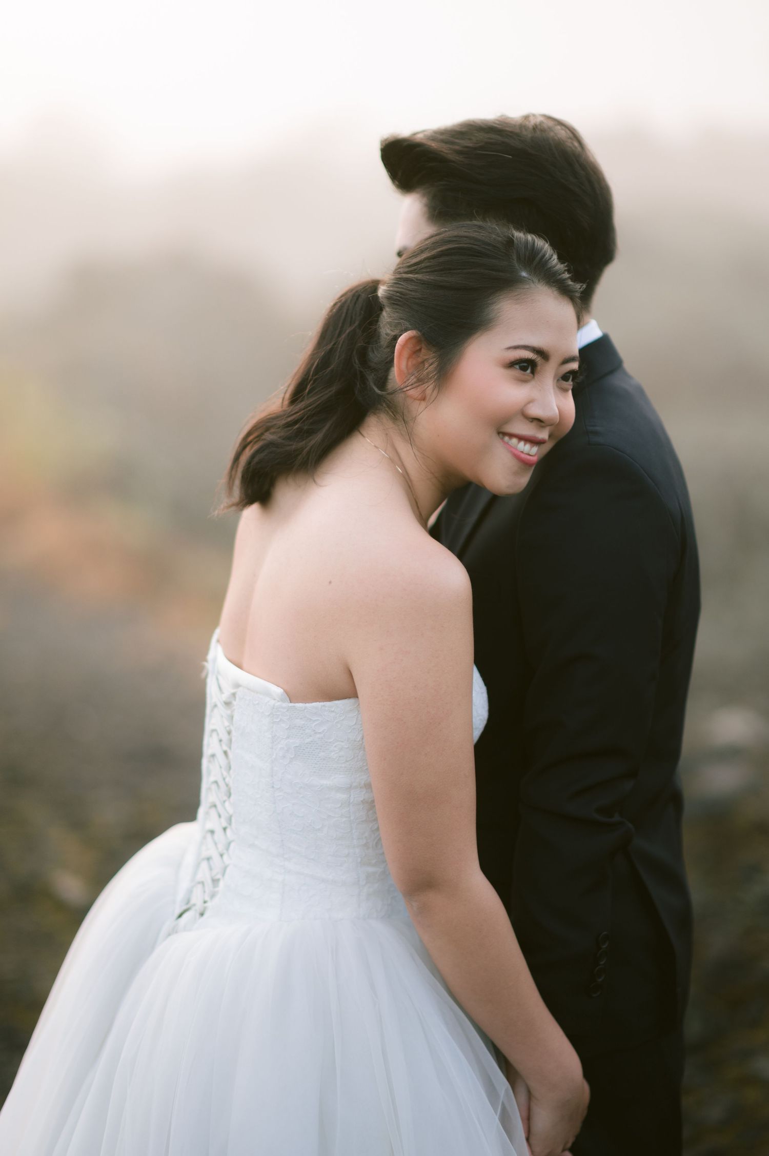Bride in white strapless gown smiles over shoulder while embracing groom in dark suit during outdoor wedding photo.