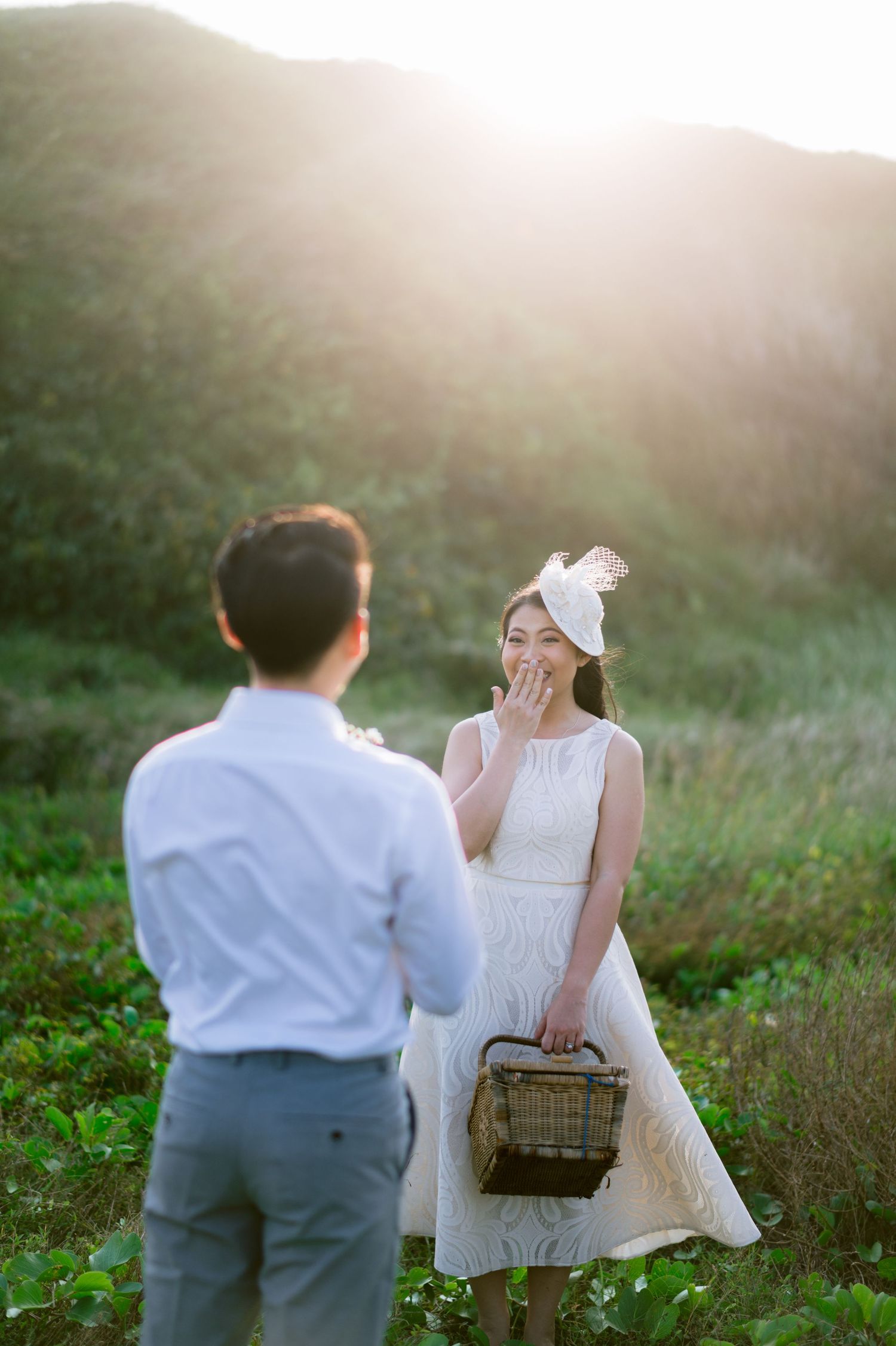 Couple sharing a romantic moment in a sunlit green field, woman in white dress and fascinator hat holding a wicker basket.