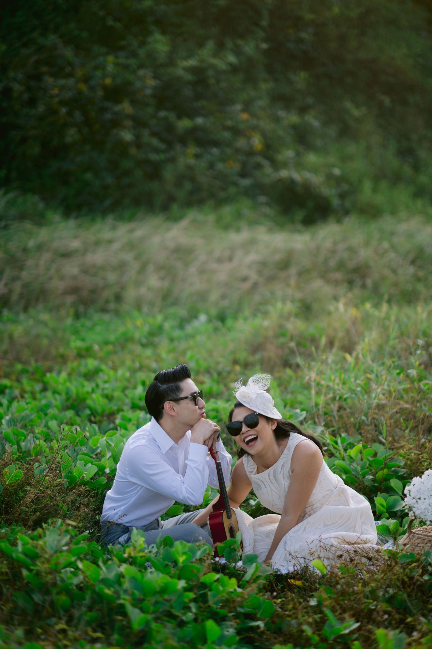 A couple sits laughing in a lush green field, dressed in white with sunglasses, sharing a romantic outdoor picnic moment.