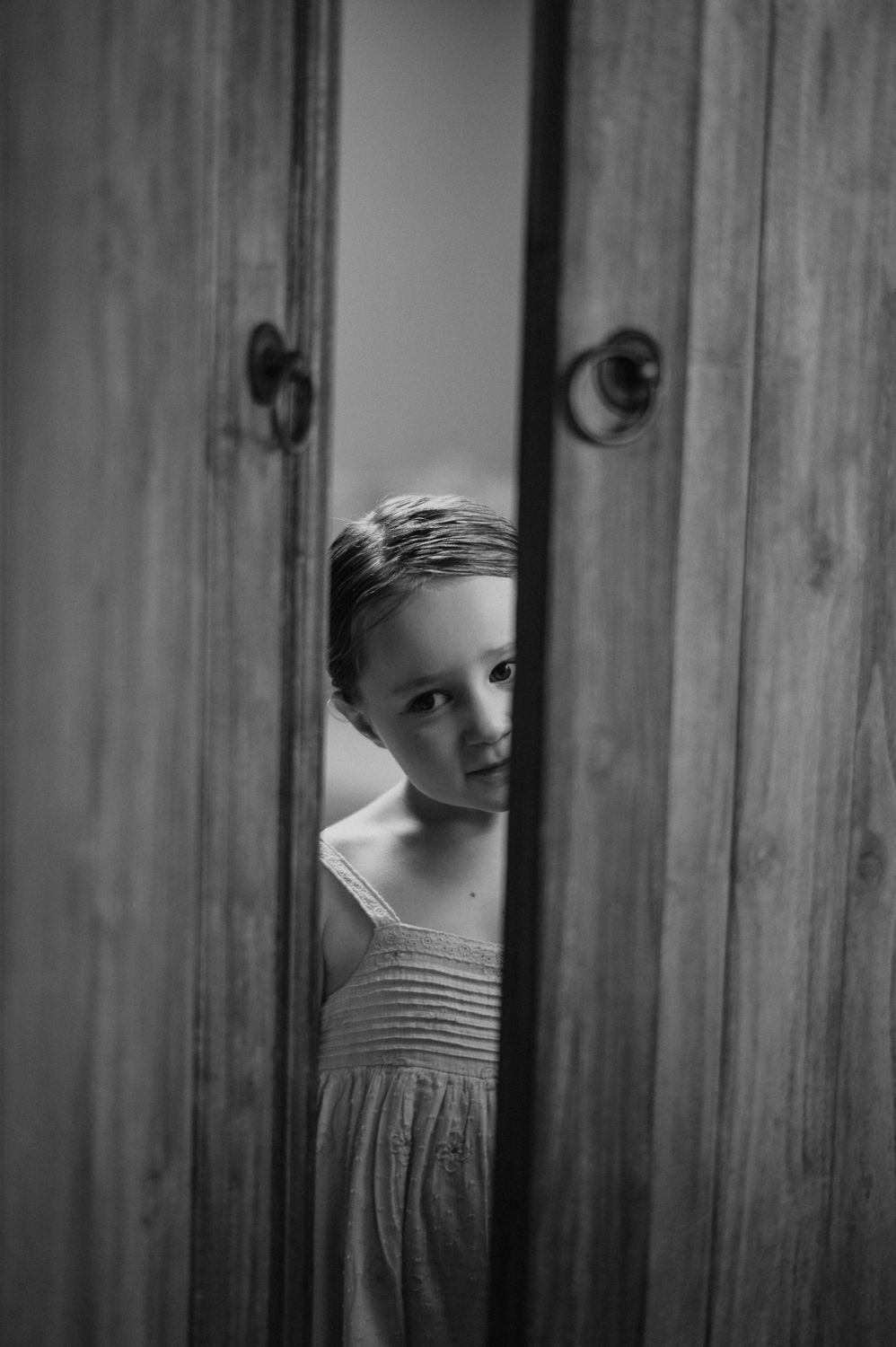 Black and white photo of a young girl peeking through wooden cabinet doors with ring handles.