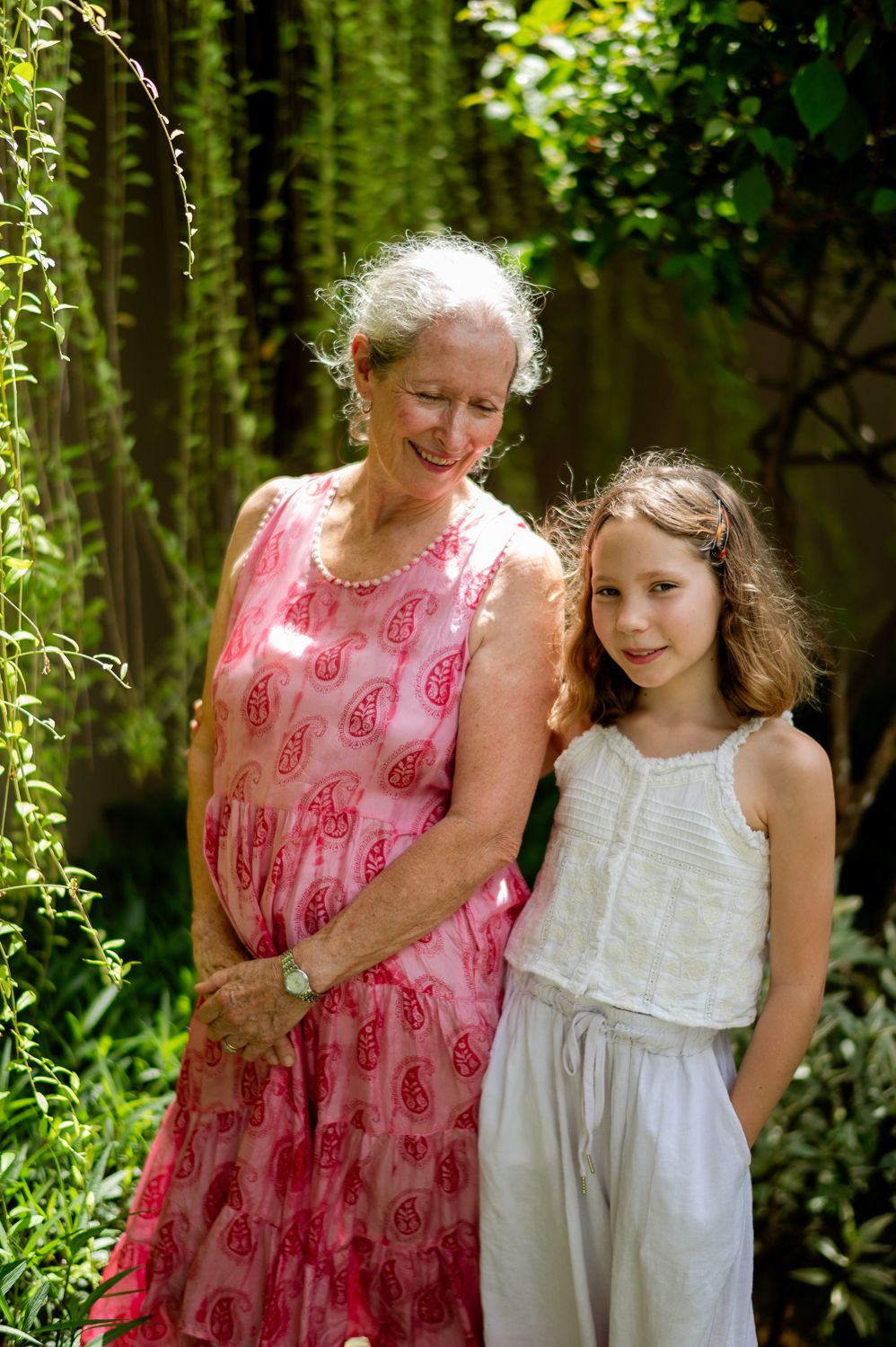 An elderly woman in a pink dress smiles beside a young girl in white outdoors in a lush green garden.