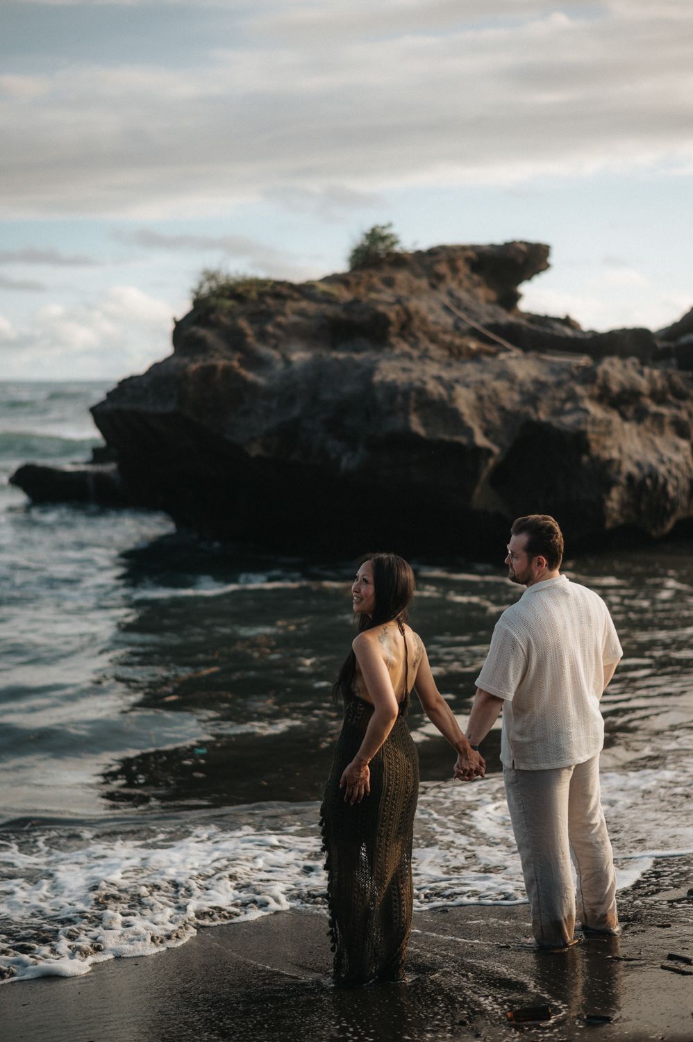 Couple holding hands at the ocean's edge with dramatic rock formations and waves at a tropical beach.