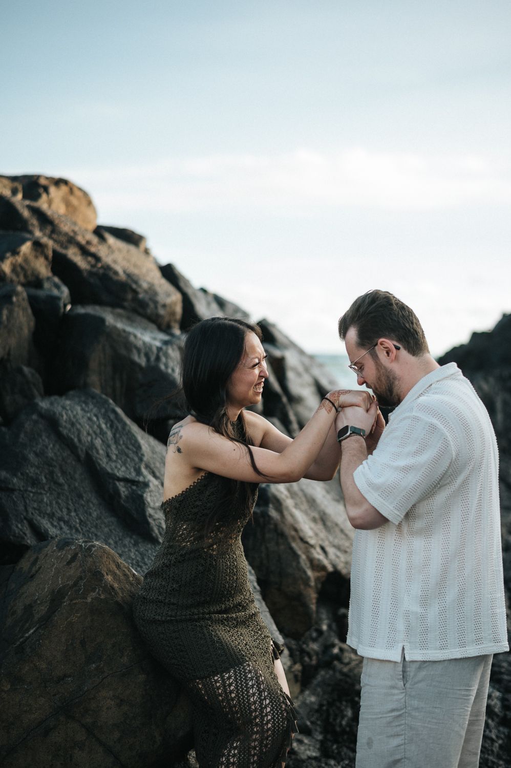 Couple holding hands romantically near rocky shore, woman in olive dress and man in white shirt, outdoor engagement photo.