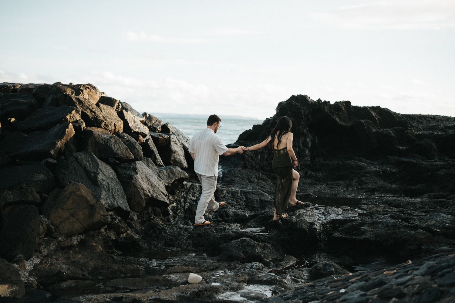 Couple holding hands while navigating rocky coastal terrain with ocean views in the background.