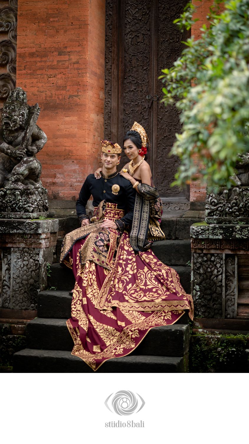 Balinese couple in traditional ceremonial attire posing on temple steps with ornate stone carvings in Bali, Indonesia.