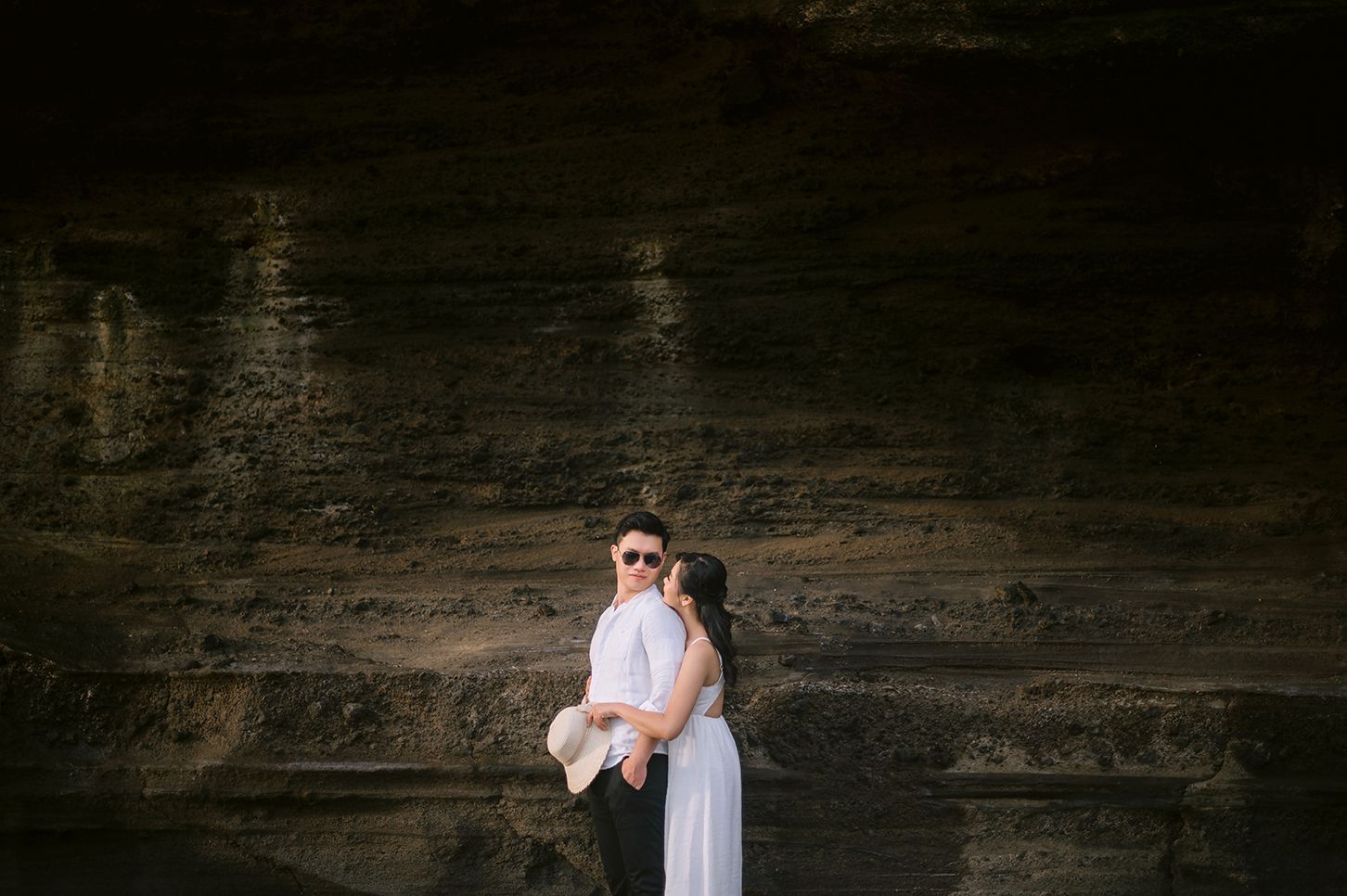 Couple embracing in front of dramatic rocky cliff wall, woman in white dress and man in dark suit.