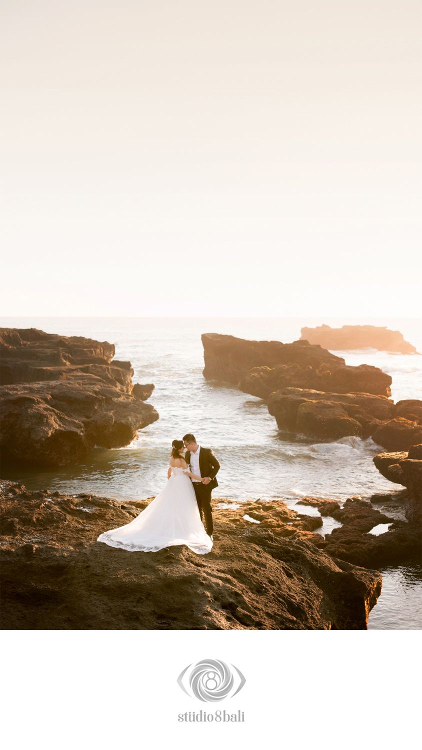 Couple sharing a romantic kiss on rocky Bali coastline at golden hour in wedding attire.