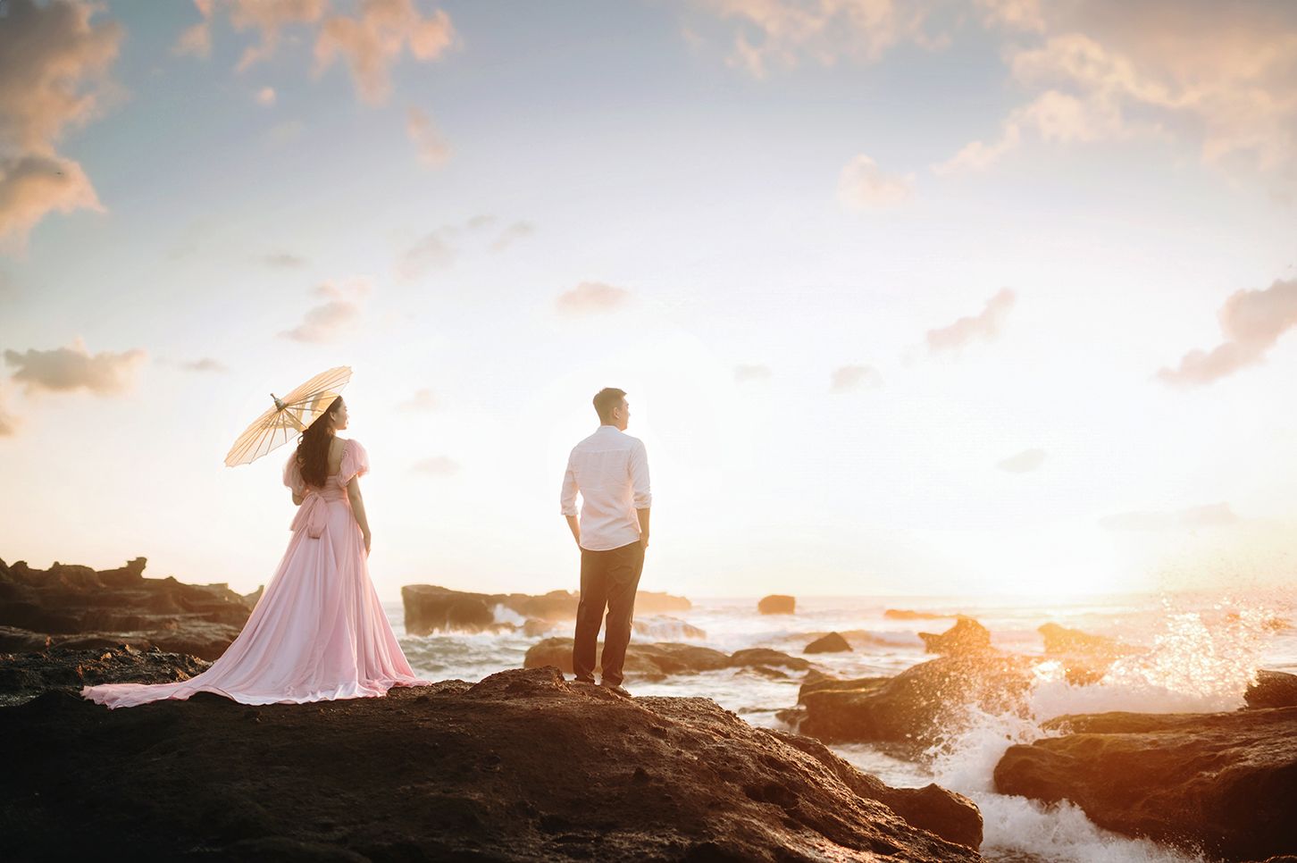 Couple standing on rocky shore at sunset, woman in pink gown with parasol, man in white shirt, ocean waves crashing nearby.
