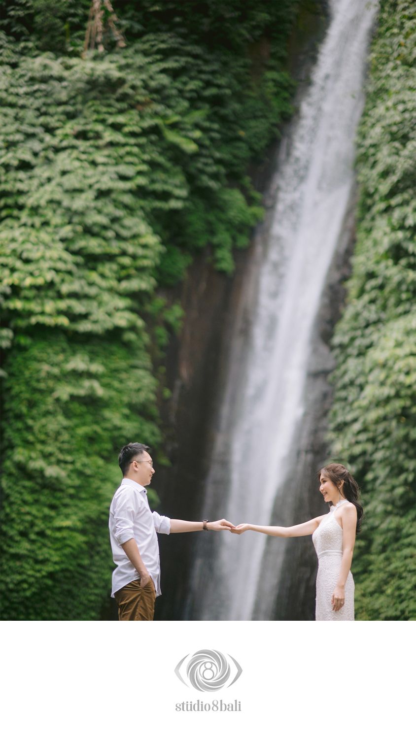 Romantic couple holding hands facing each other before a stunning green waterfall backdrop in Bali.