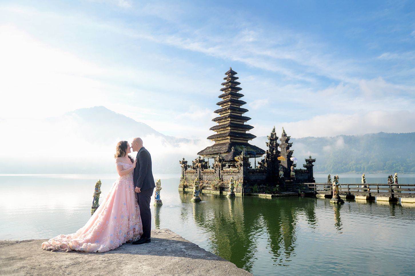 Couple in wedding attire poses before Pura Ulun Danu Bratan temple on Bali lake with misty mountain backdrop.