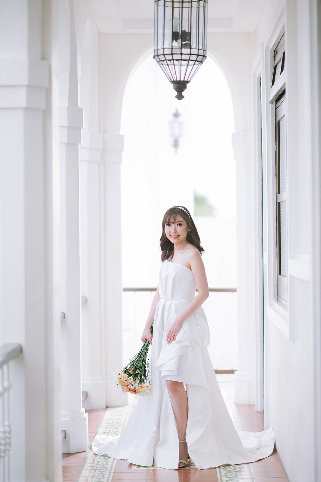 A bride in a white strapless gown holds yellow flowers in an elegant arched corridor with pendant lights.