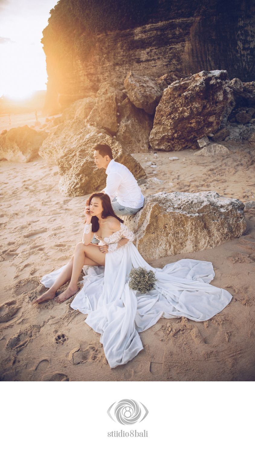 Couple in white wedding attire posing on sandy beach at sunset with dramatic rocky cliffs in Bali.