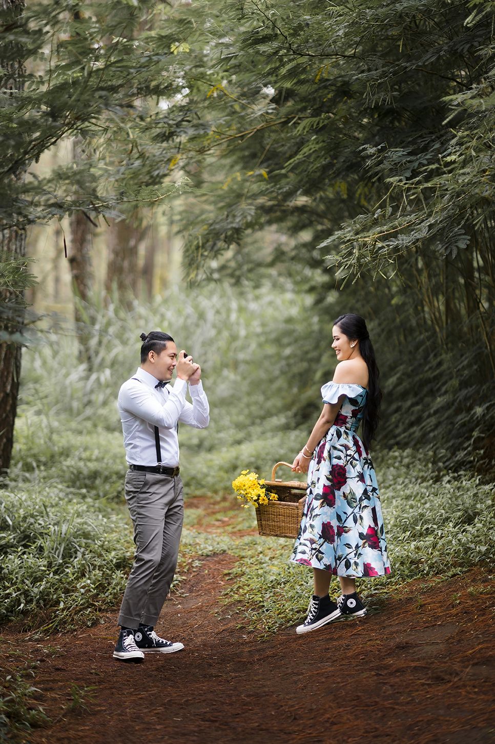A man photographs a smiling woman in a floral dress holding a basket of yellow flowers on a forest path.