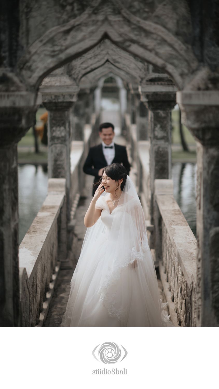 Bride in white gown and veil stands on stone bridge with groom in tuxedo at elegant Bali wedding venue.