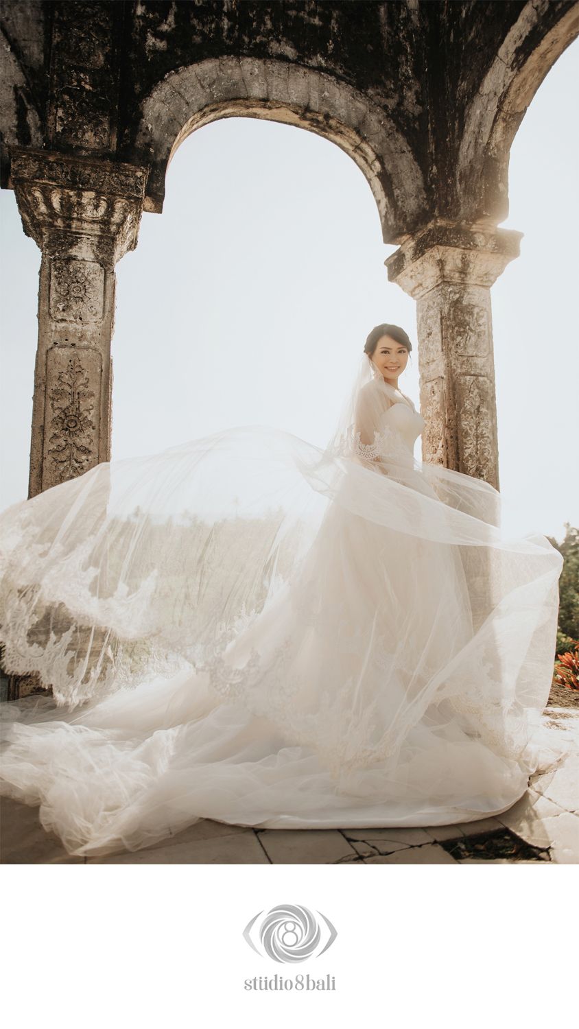 Bride in flowing white wedding gown with veil billowing under stone archway at Bali venue, Studio 8 Bali photography.