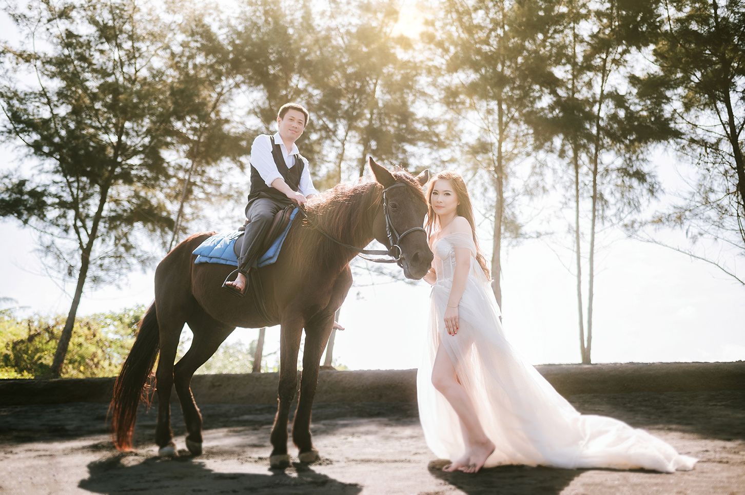 Groom on horseback beside bride in flowing gown during golden sunset wedding photo shoot outdoors.