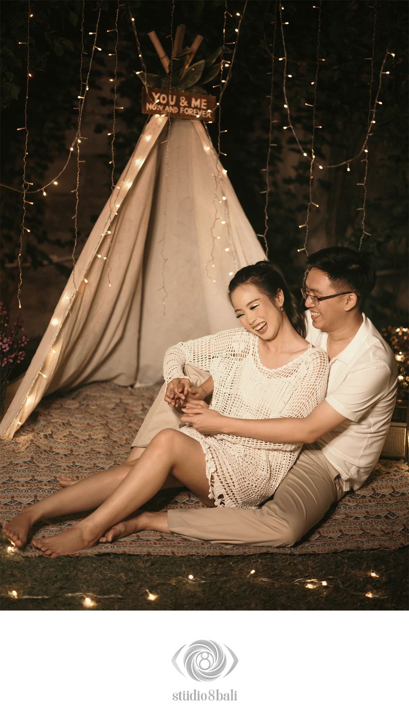 Couple sitting together by a boho teepee with fairy lights, woman in crochet dress, romantic outdoor night setting.
