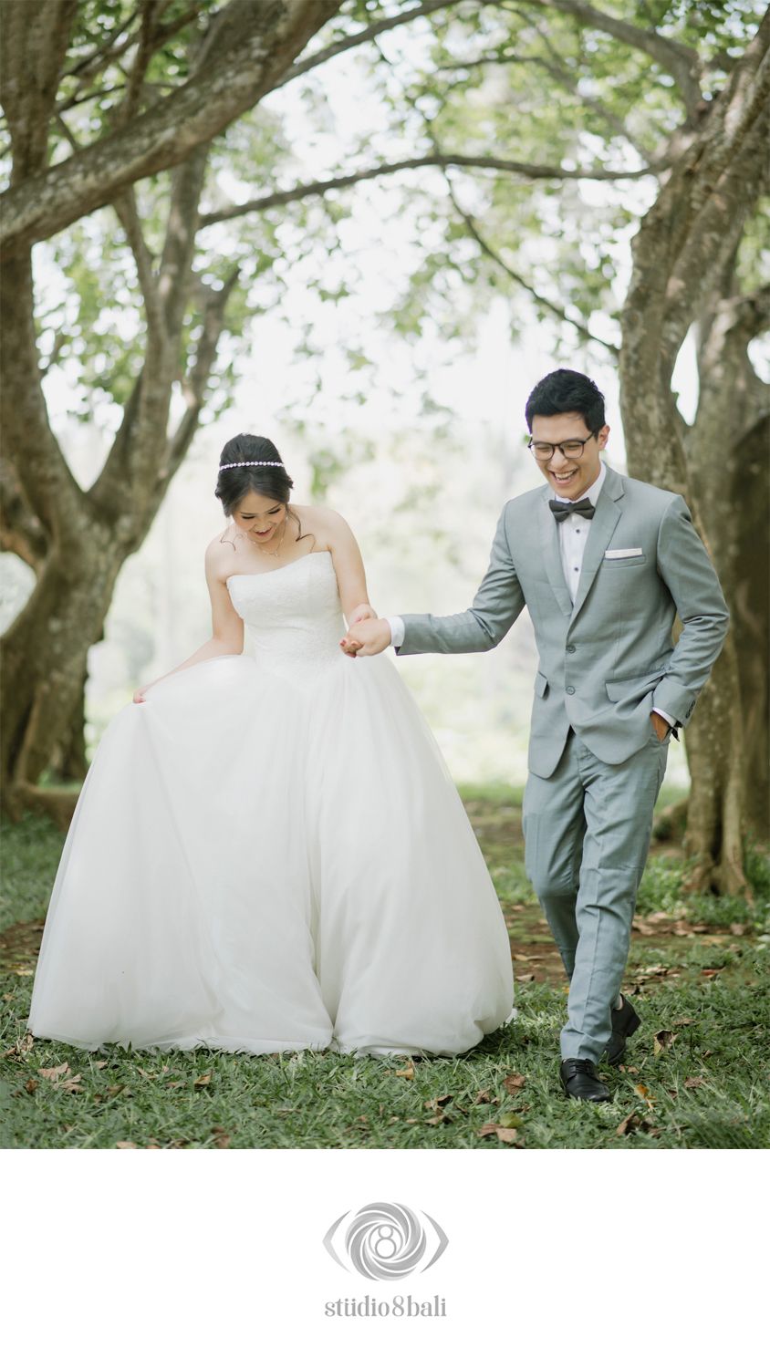 Bride in white ball gown and groom in gray suit walk hand-in-hand through a tree-lined garden path.
