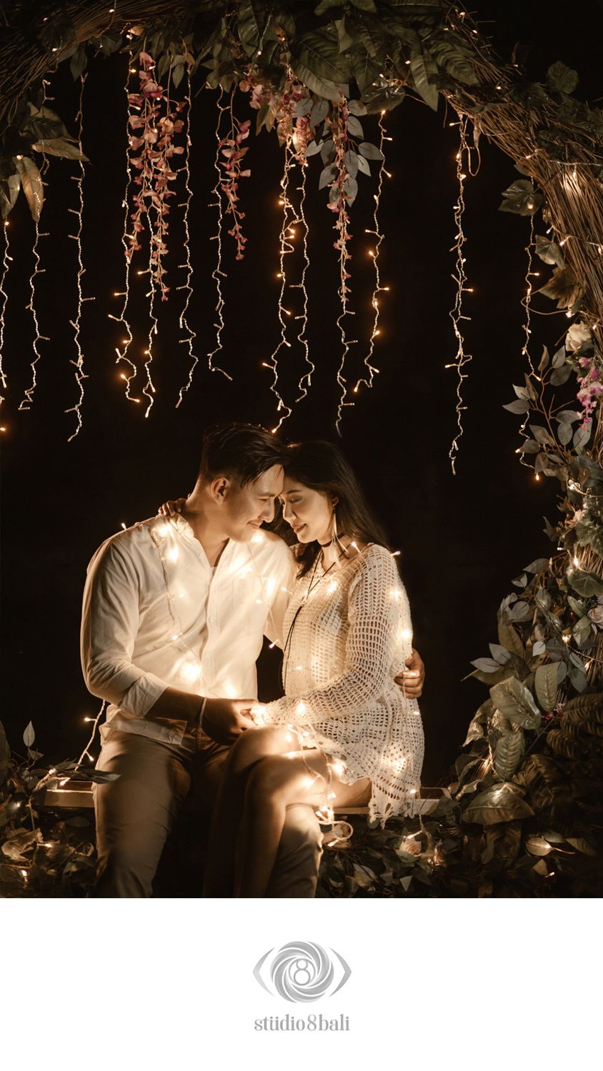 Romantic couple sitting under fairy lights and floral arch in a warm, intimate engagement photo by Studio 8 Bali.