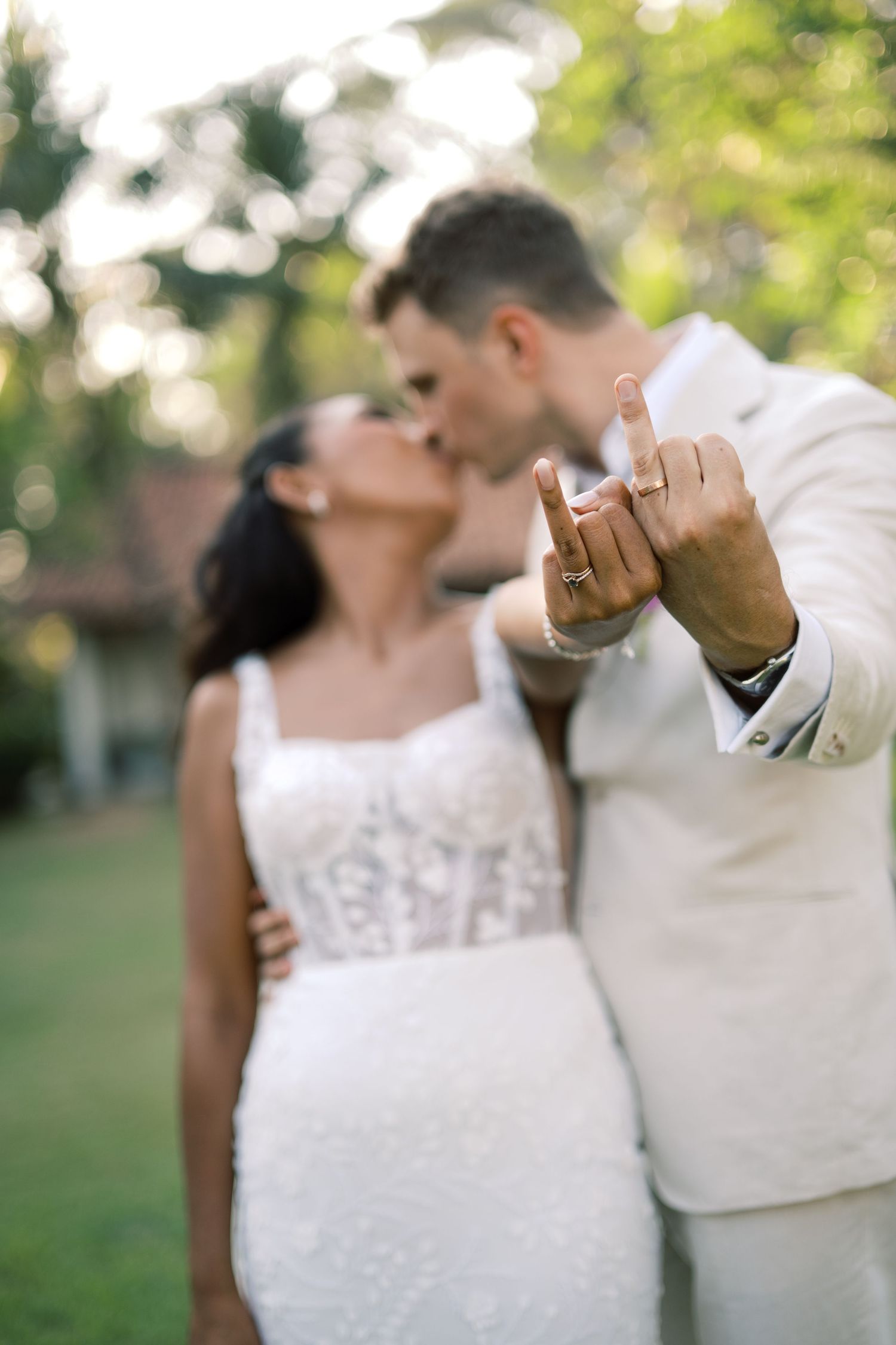Newlyweds kiss while groom displays wedding ring to camera in a romantic outdoor garden setting.