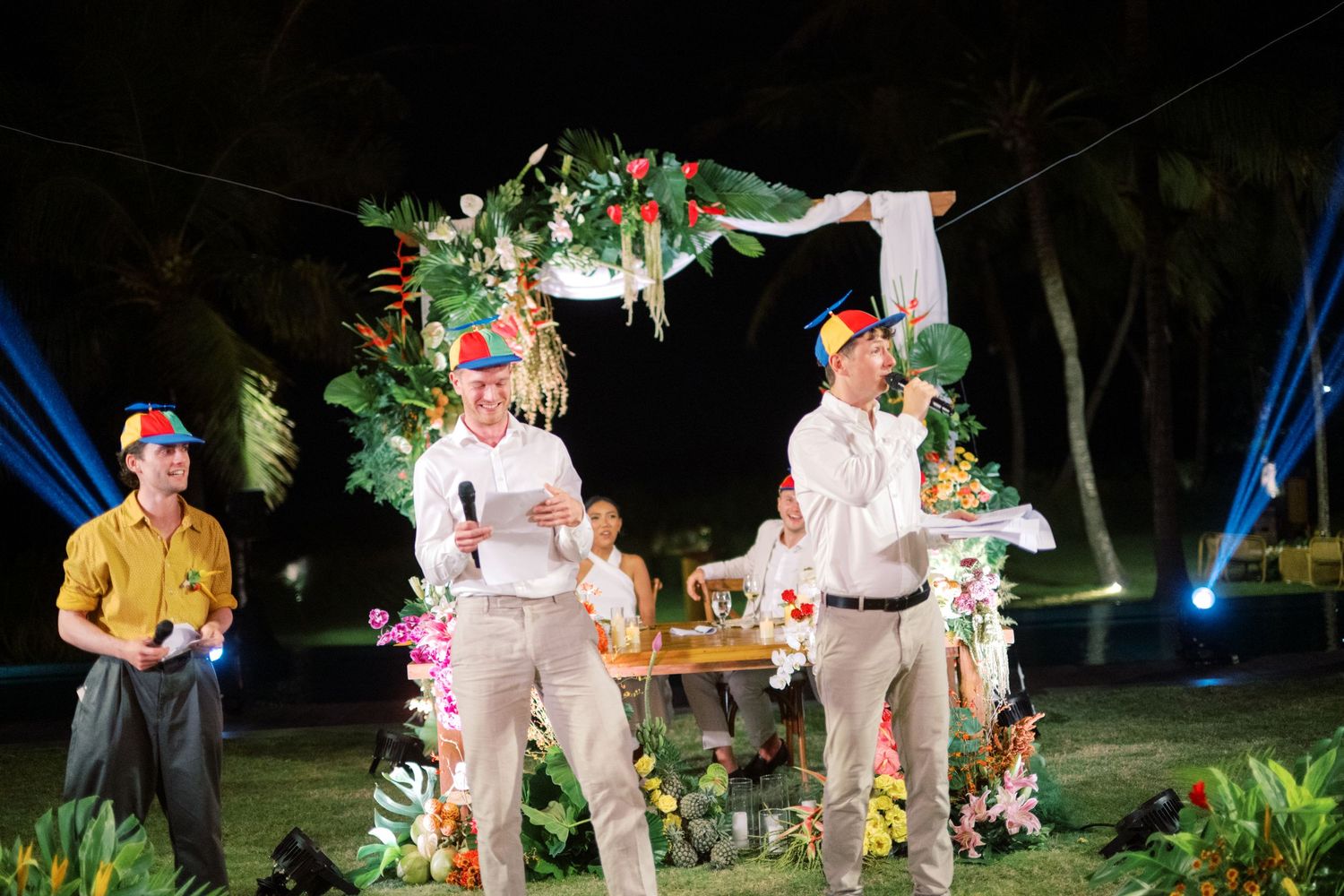 Two men in colorful hats speak at decorated outdoor wedding reception stage at night.