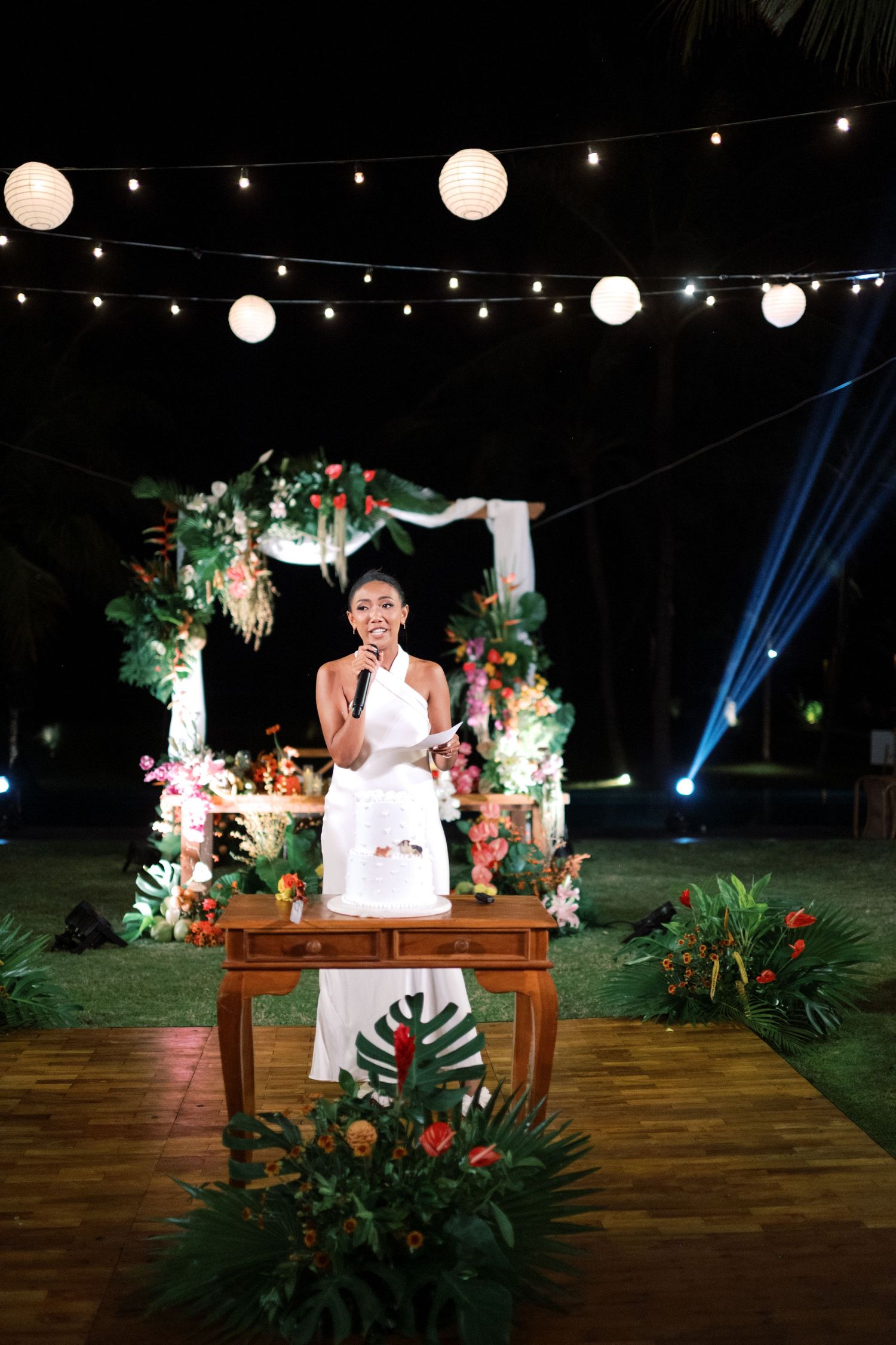Woman in white dress gives speech at outdoor tropical wedding reception under paper lantern lights.
