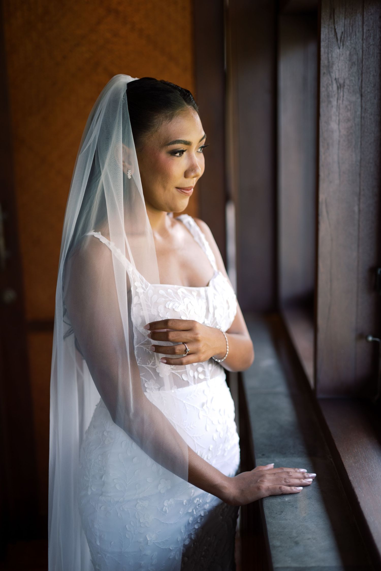 Beautiful bride in lace wedding gown and veil gazes out window in soft natural light.
