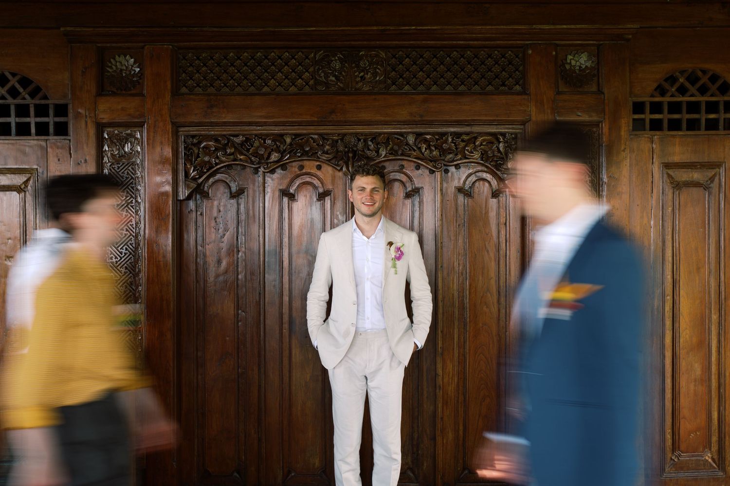 Groom in beige suit stands confidently before ornate wooden wall with blurred guests passing.