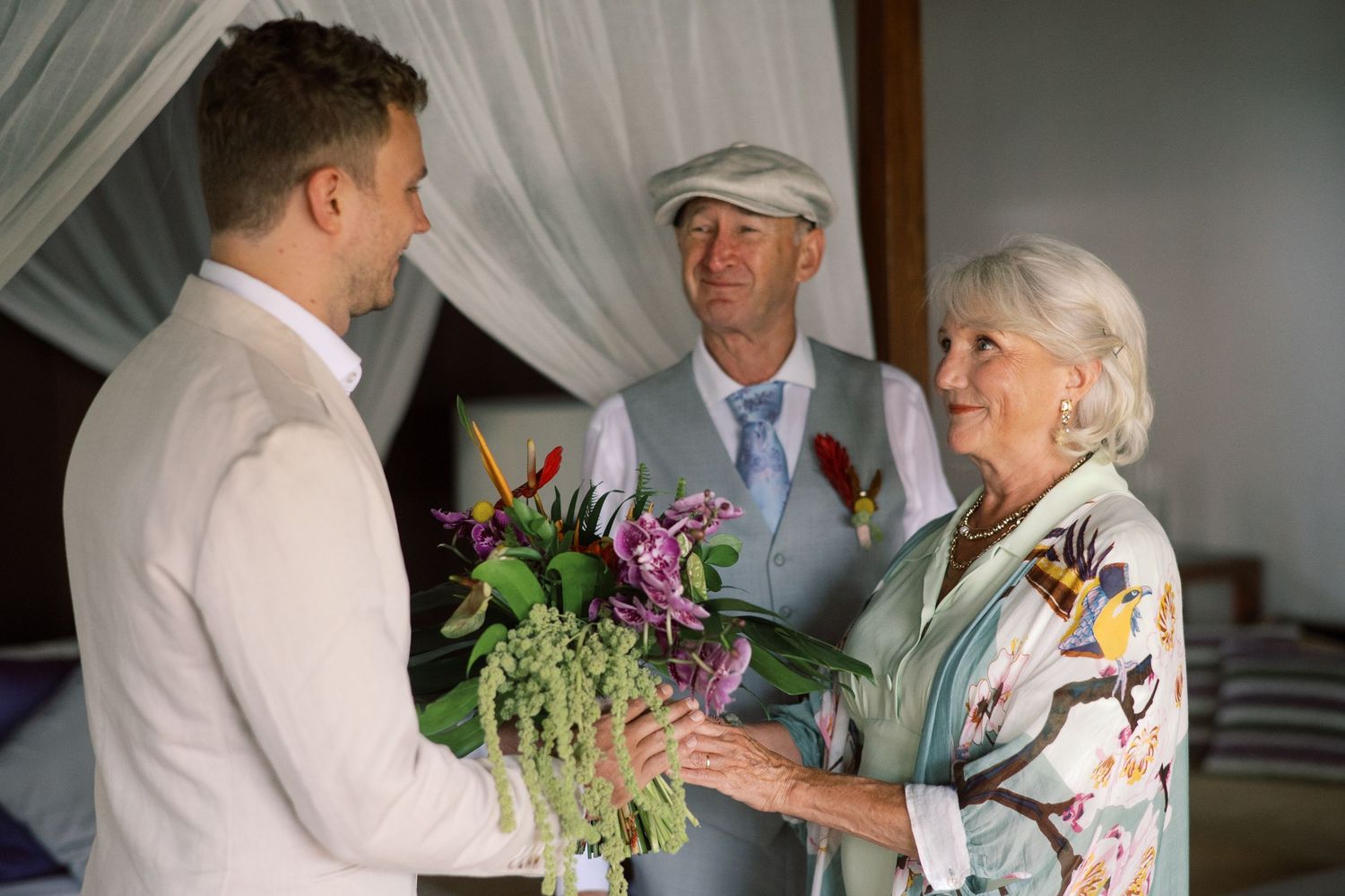 Elegant wedding scene with groom receiving flowers from older woman, officiant in flat cap standing behind them.