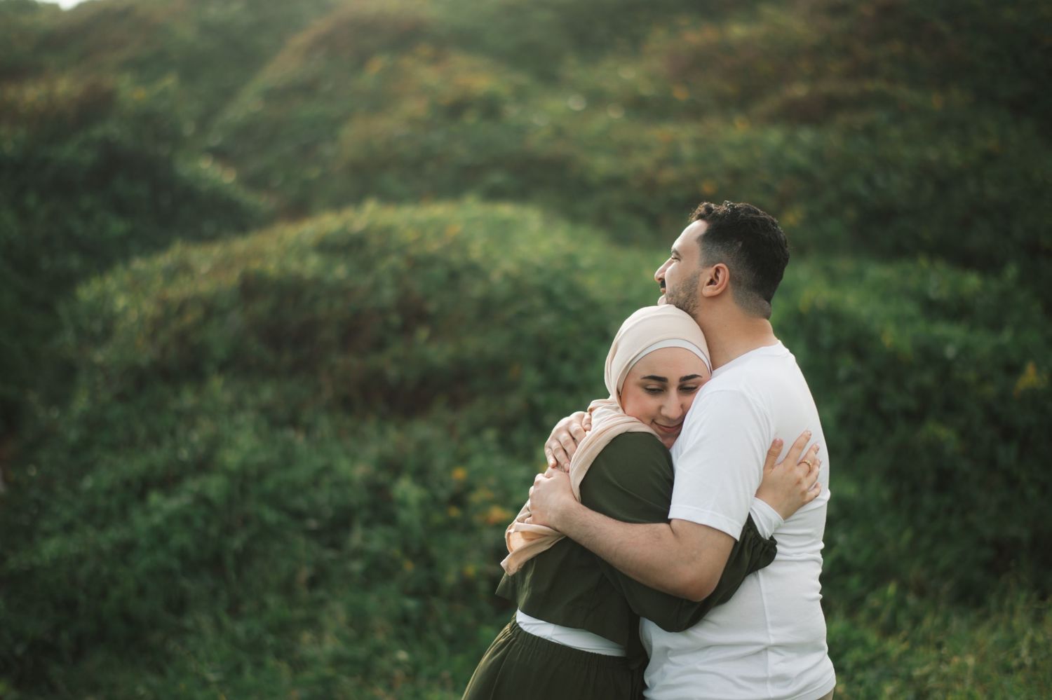 A couple embracing outdoors in a lush green field, woman wearing a hijab, sharing a tender moment.