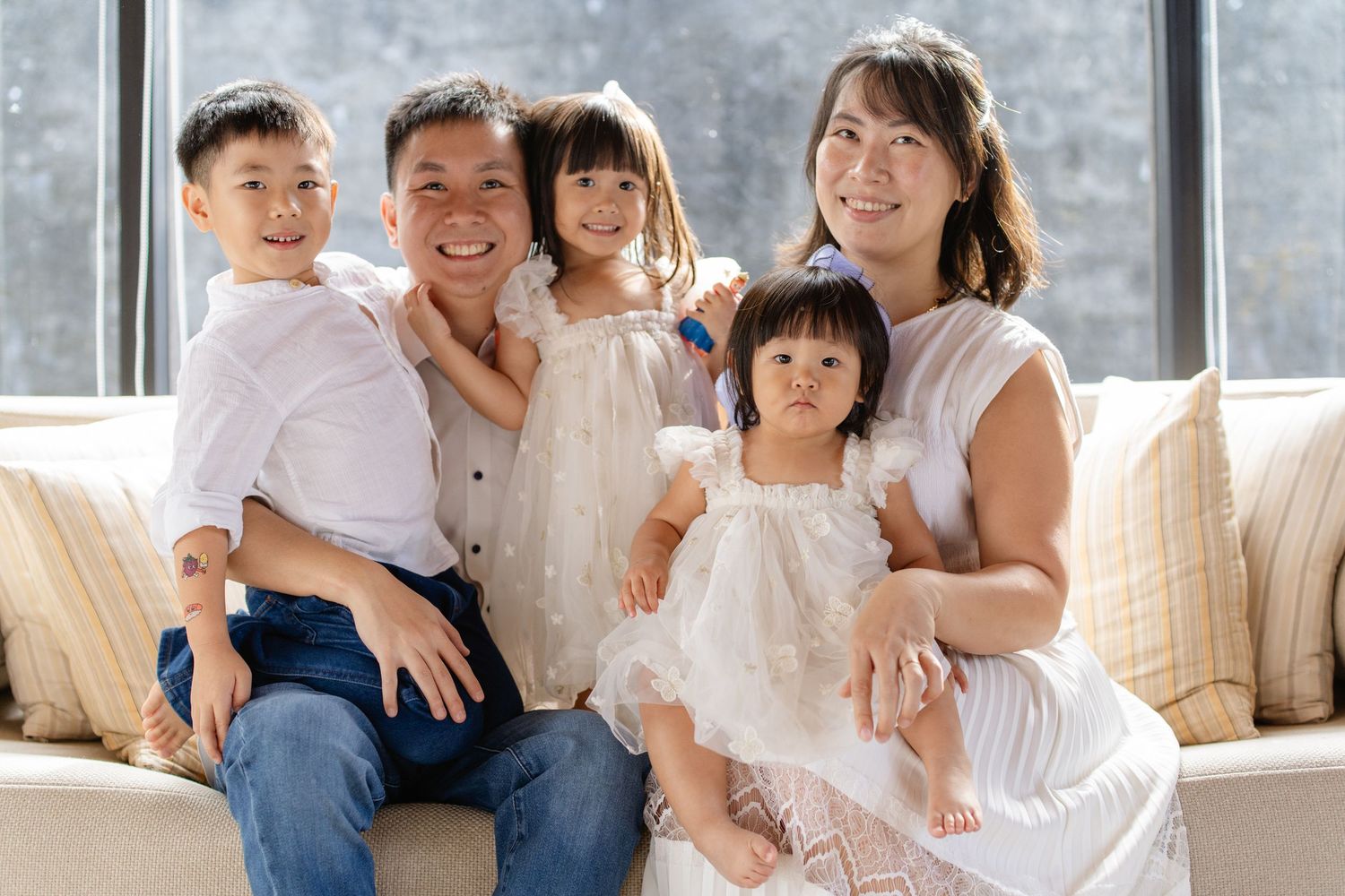 Happy Asian family of five posing together on a couch, smiling warmly in a bright, elegant living room setting.