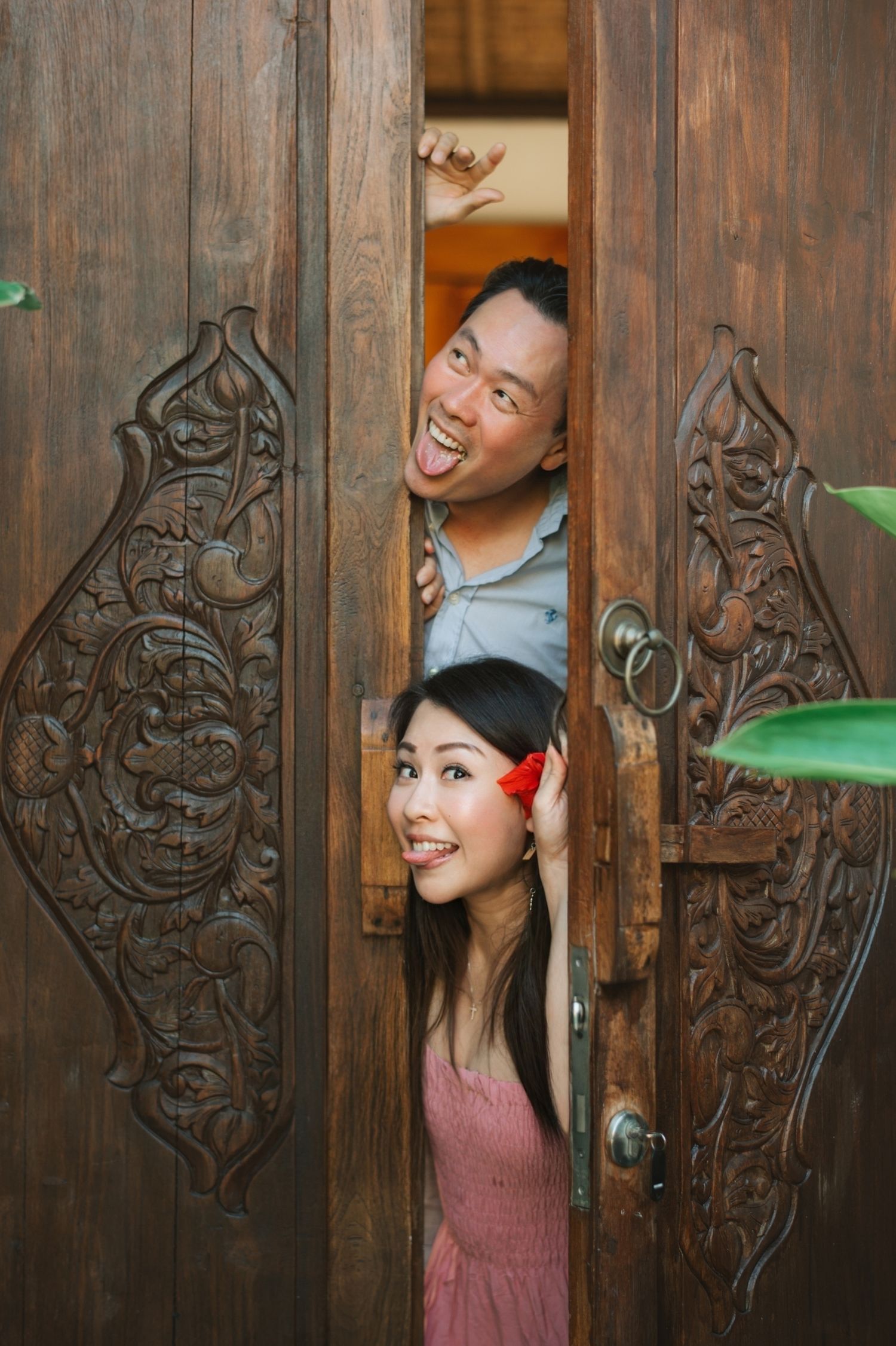 A joyful Asian couple peeking through ornately carved wooden doors, smiling and laughing playfully.