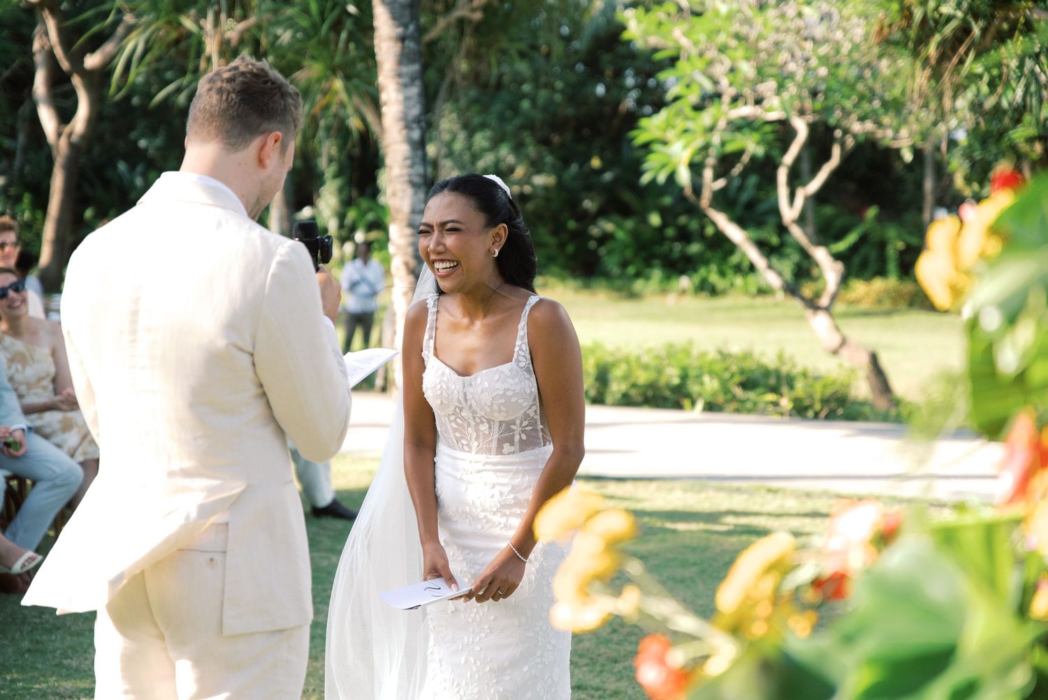 Bride in white lace gown laughing joyfully with groom in cream suit during outdoor garden wedding ceremony.